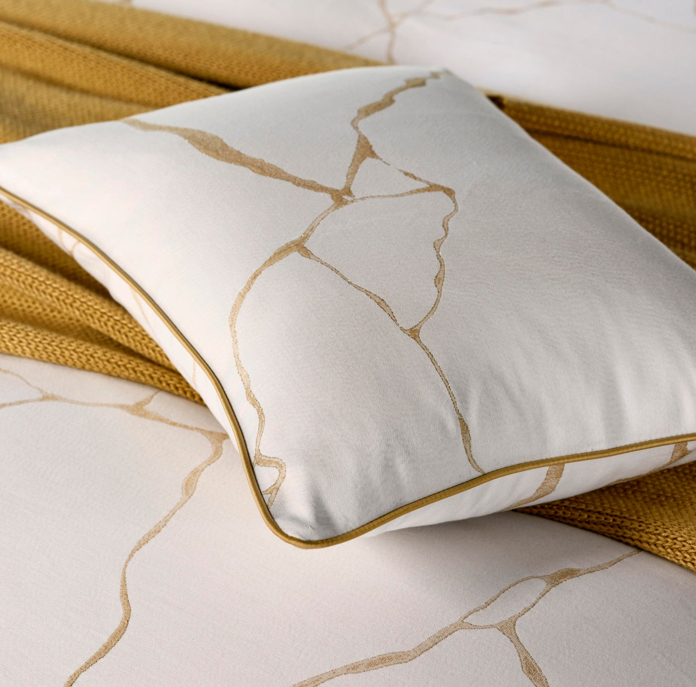A white throw pillow with a golden crackle pattern and piping rests on a textured golden throw. featuring Jessie Pillowcase by Celso de Lemos available at Linen Society.