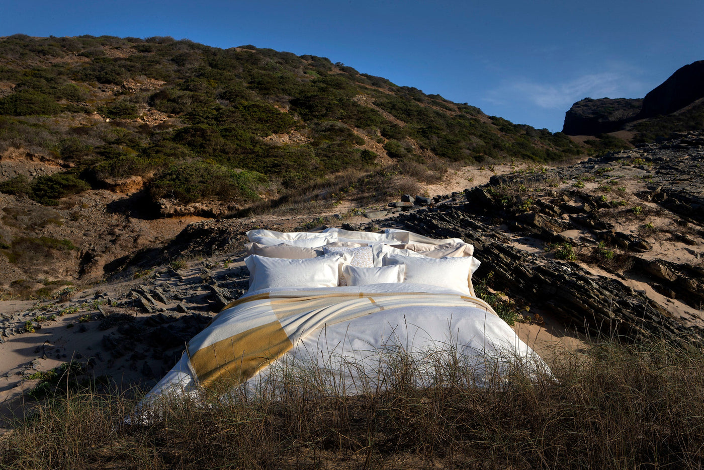 A bed is set up outdoors on a sandy, rocky landscape with hills in the background, featuring the Joanne Pillowcase by Celso de Lemos available at Linen Society.