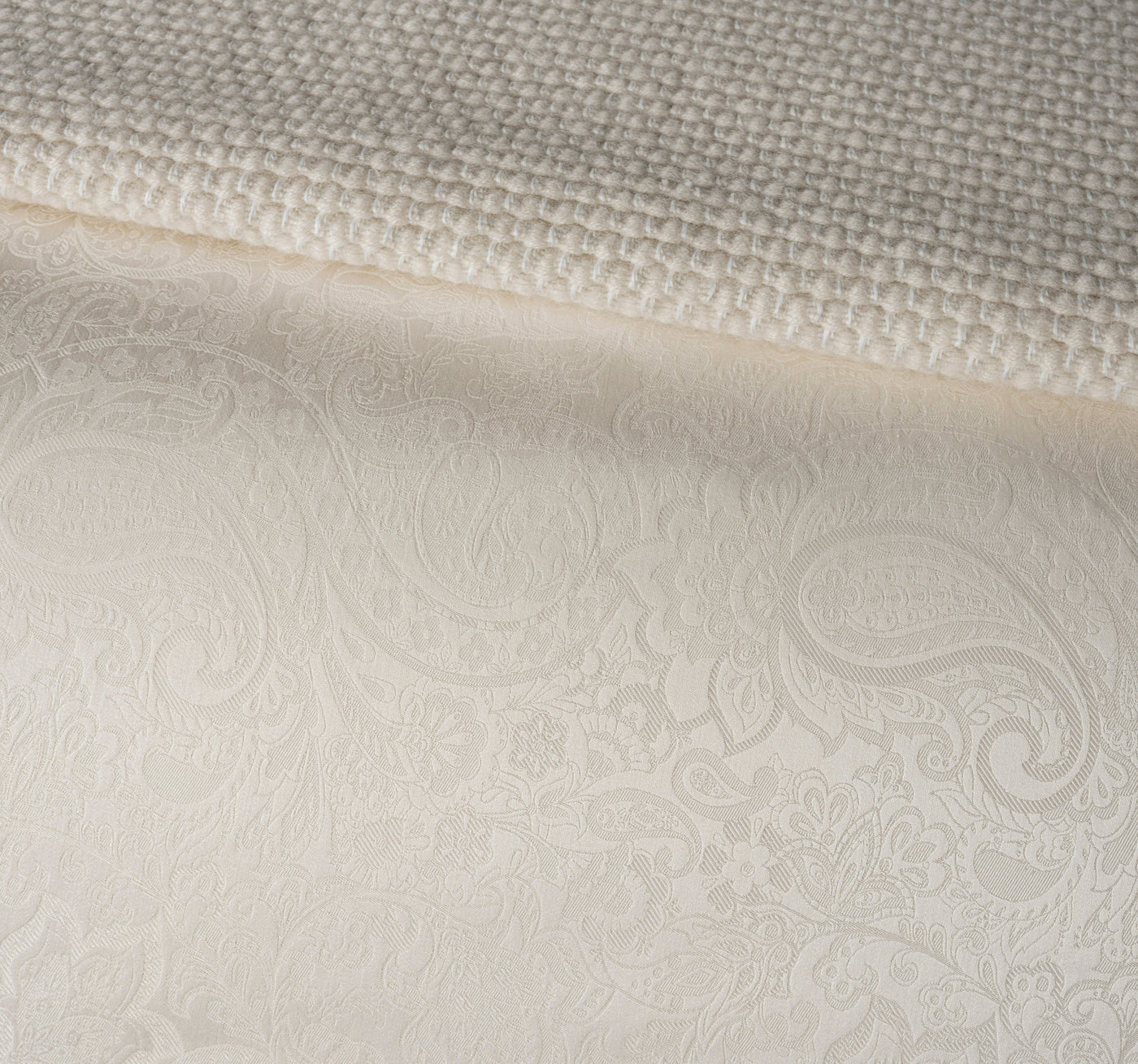 A close-up view of a textured, woven fabric laid over a patterned fabric featuring embossed paisley motifs. featuring Joanne Pillowcase by Celso de Lemos available at Linen Society.