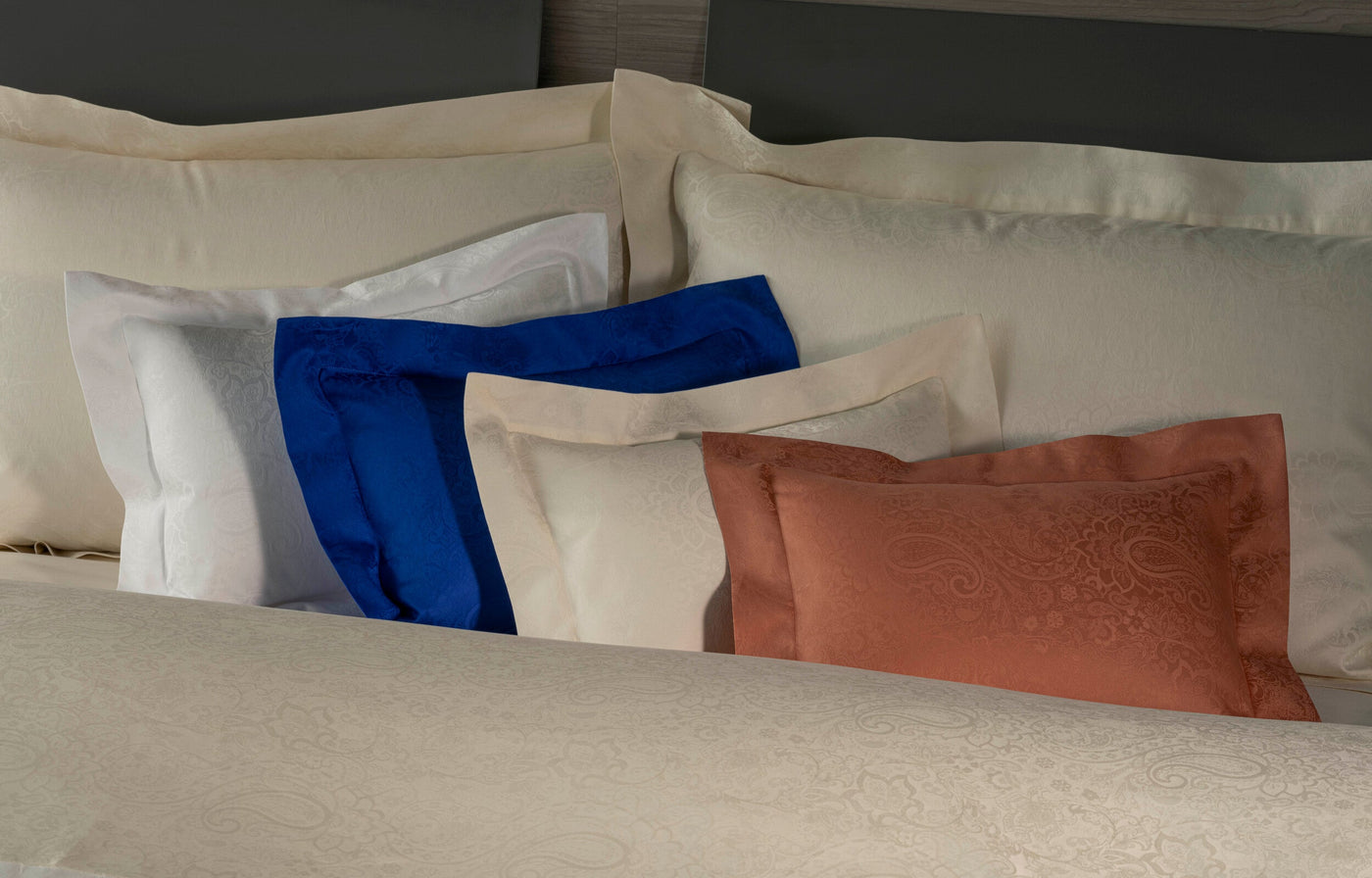 Multiple pillowcases with a subtle paisley damask pattern in cream, white, blue, and rust colors, featuring Joanne Pillowcase by Celso de Lemos available at Linen Society.