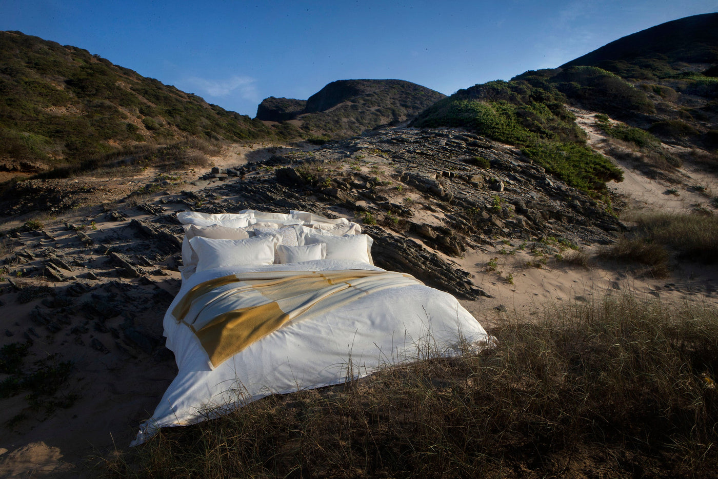 A bed dressed in white linens and a gold-accented throw is set against a rugged, coastal landscape. featuring Joanne Pillowcase by Celso de Lemos available at Linen Society.