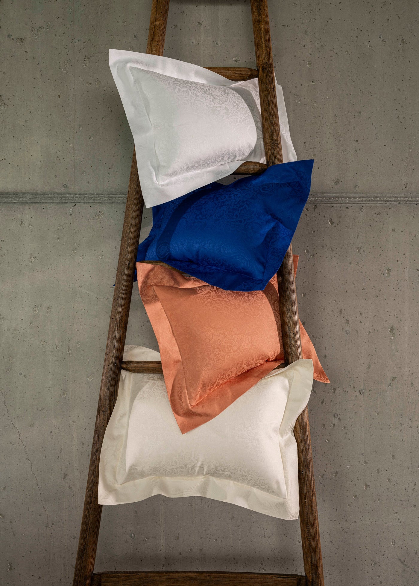 Four patterned pillowcases in white, blue, and coral are stacked on a wooden ladder against a concrete wall, featuring Joanne Pillowcase by Celso de Lemos available at Linen Society.