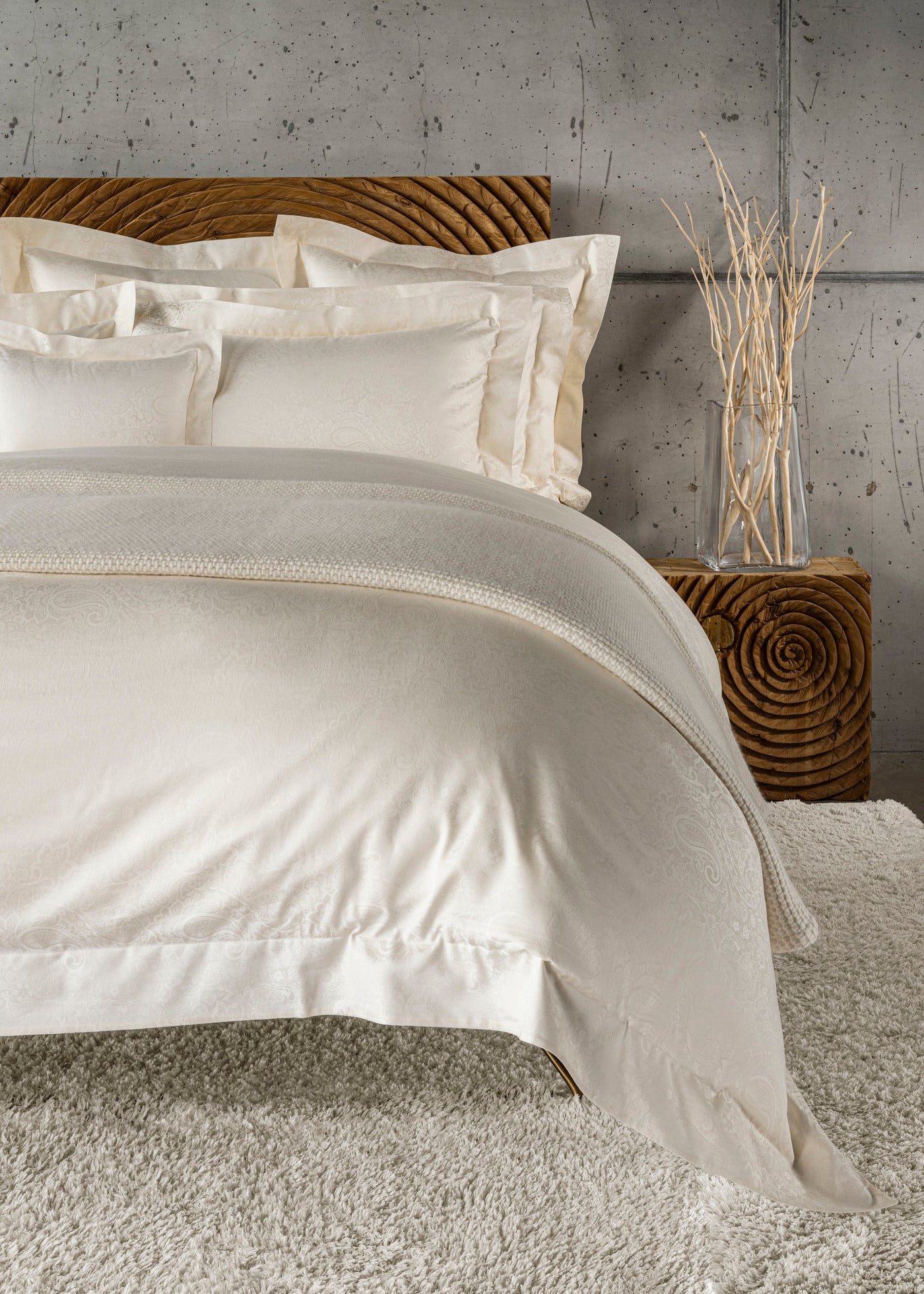 A bed features neatly folded, off-white pillowcases and a duvet with a subtle paisley pattern, laid out on a plush rug next to a textured wooden nightstand with dried branches in a glass vase, featuring Joanne Pillowcase by Celso de Lemos available at Linen Society.