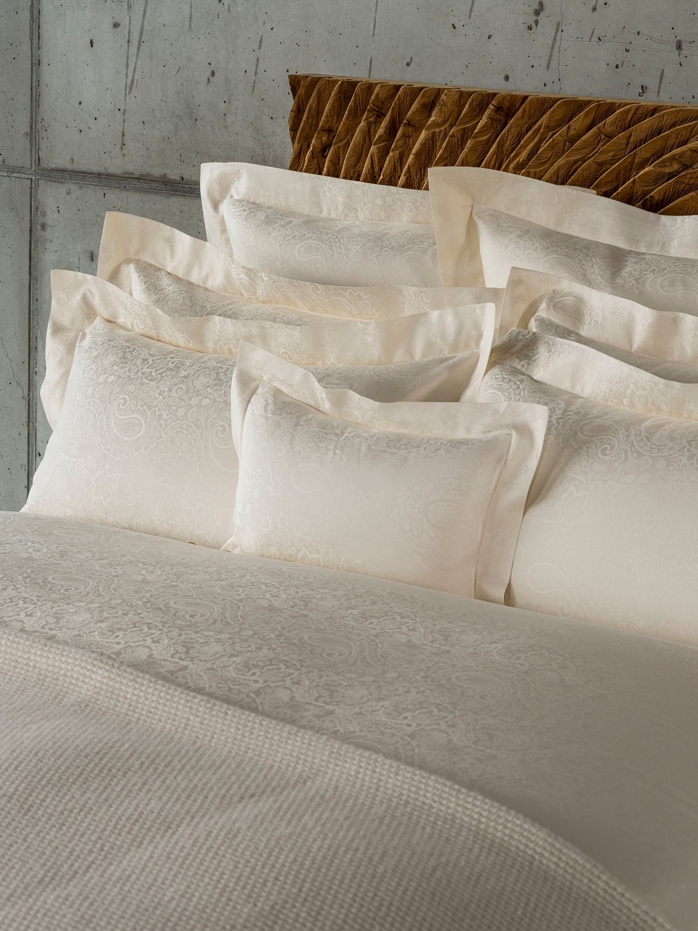White paisley pattern accent pillows are stacked on a bed with a textured headboard, featuring Joanne Pillowcases by Celso de Lemos available at Linen Society.