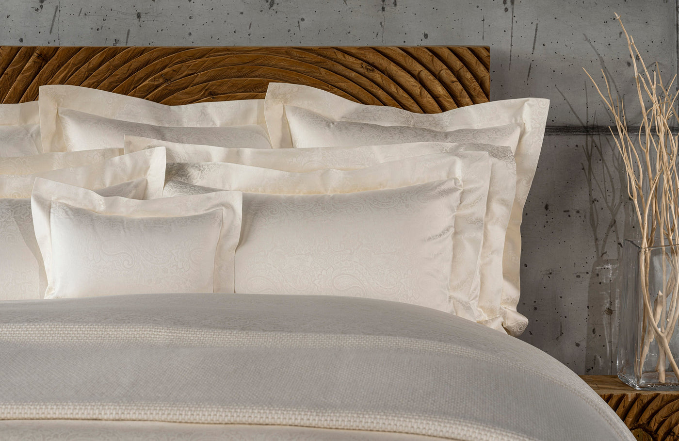 Stacked ivory Pillowcases with subtle paisley pattern displayed on a bed with a textured wooden headboard, featuring Joanne Pillowcase by Celso de Lemos available at Linen Society.