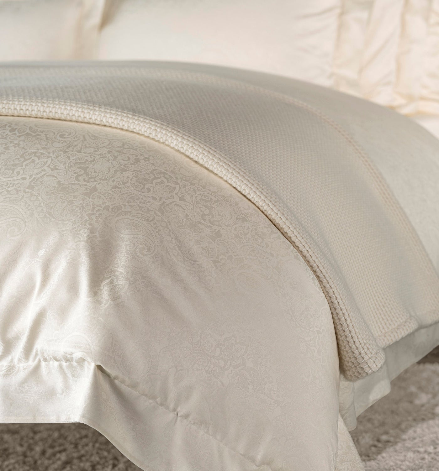 Close-up of a cream duvet cover with a subtle paisley pattern and a textured knitted throw, featuring Joanne Pillowcase by Celso de Lemos available at Linen Society.