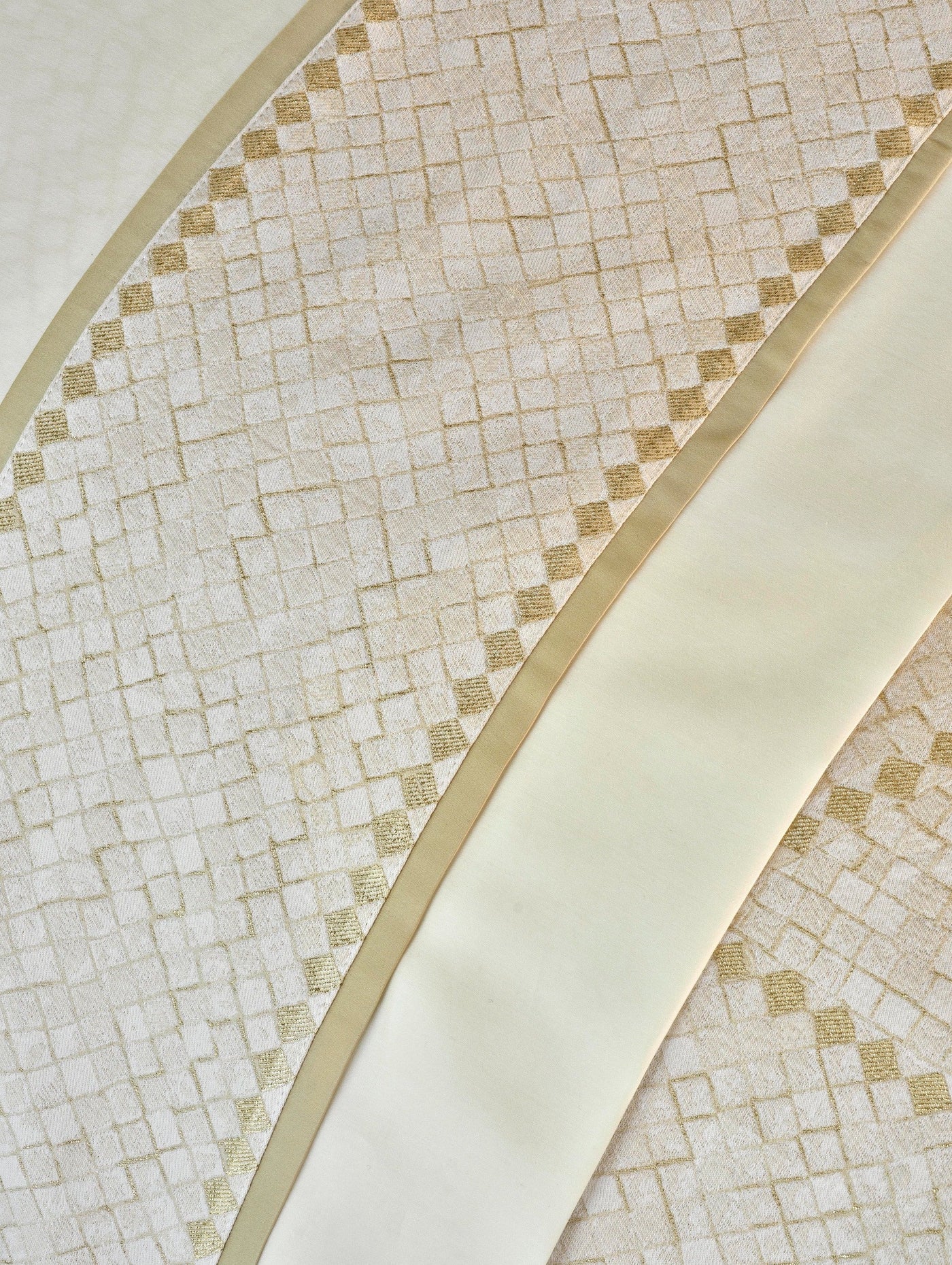 Close-up of cream fabric with a textured geometric pattern and a subtle gold thread, accented by a smooth satin band. featuring Kerylos Pillowcase by Celso de Lemos available at Linen Society.