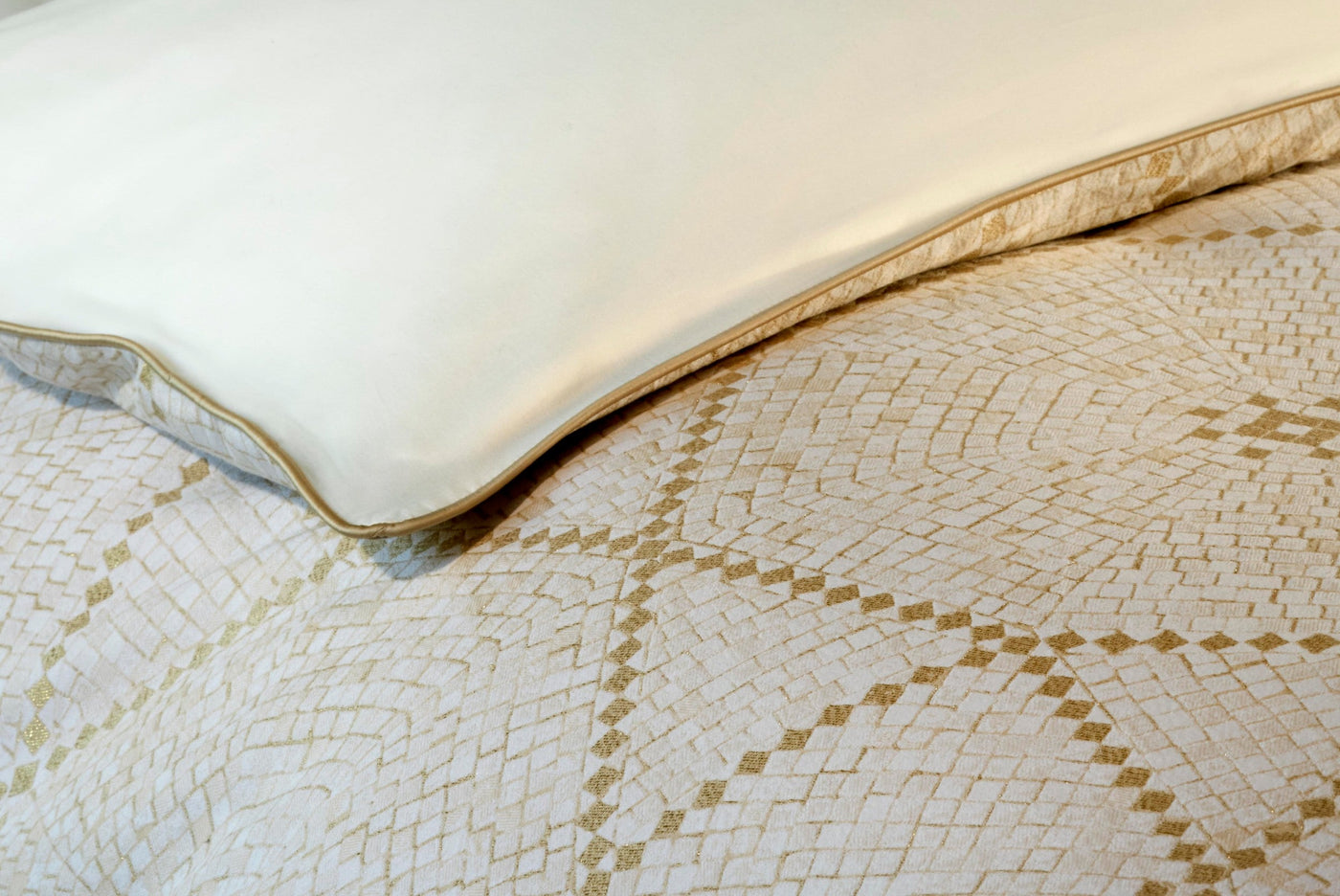 Close-up of a cream pillow with a gold trim resting on a textured fabric with a gold geometric pattern, featuring Kerylos Pillowcase by Celso de Lemos available at Linen Society.