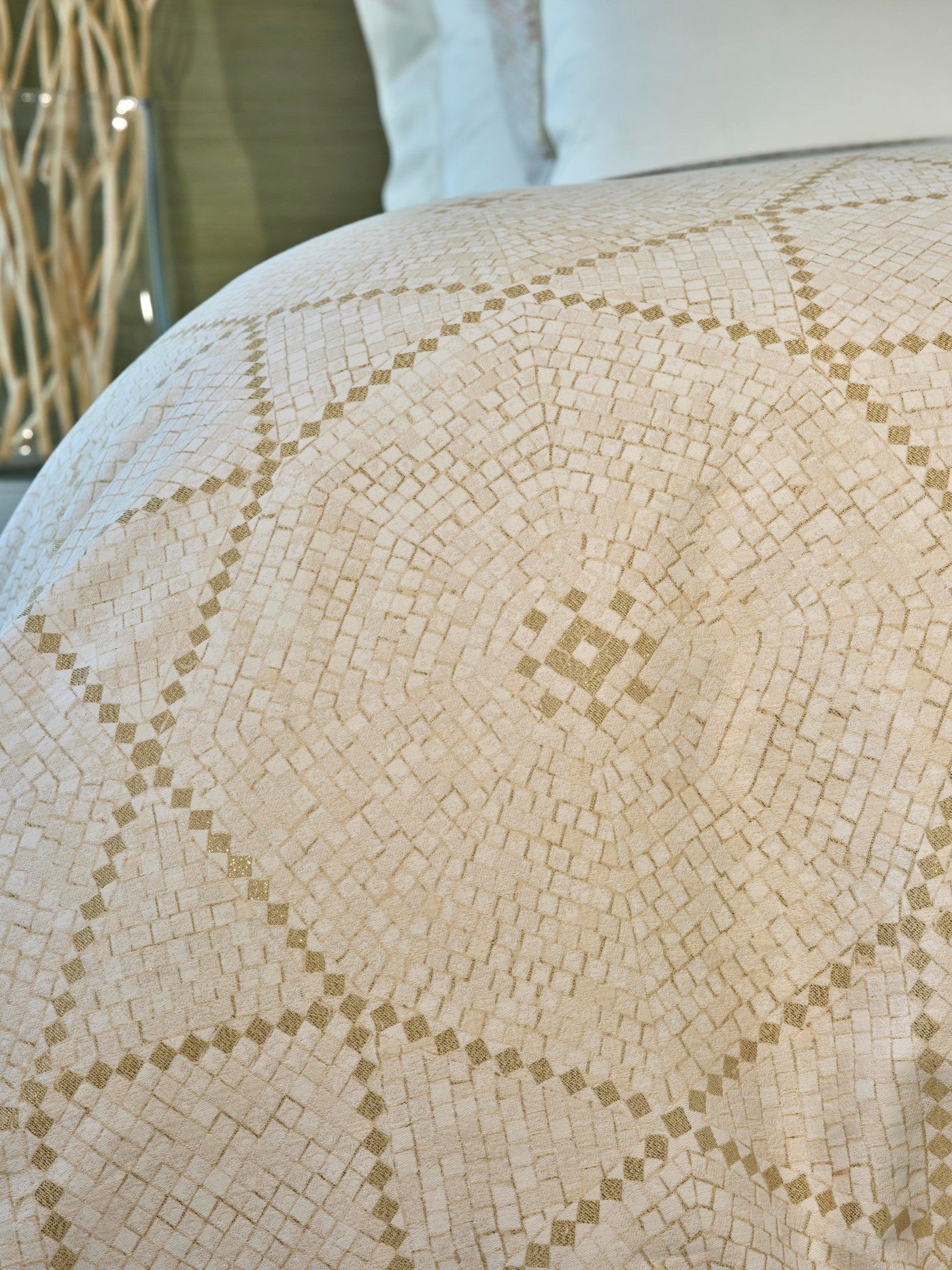 A neutral-toned pillowcase with a subtle mosaic-like pattern featuring shimmering gold accents, draped on a bed featuring Kerylos Pillowcase by Celso de Lemos available at Linen Society.