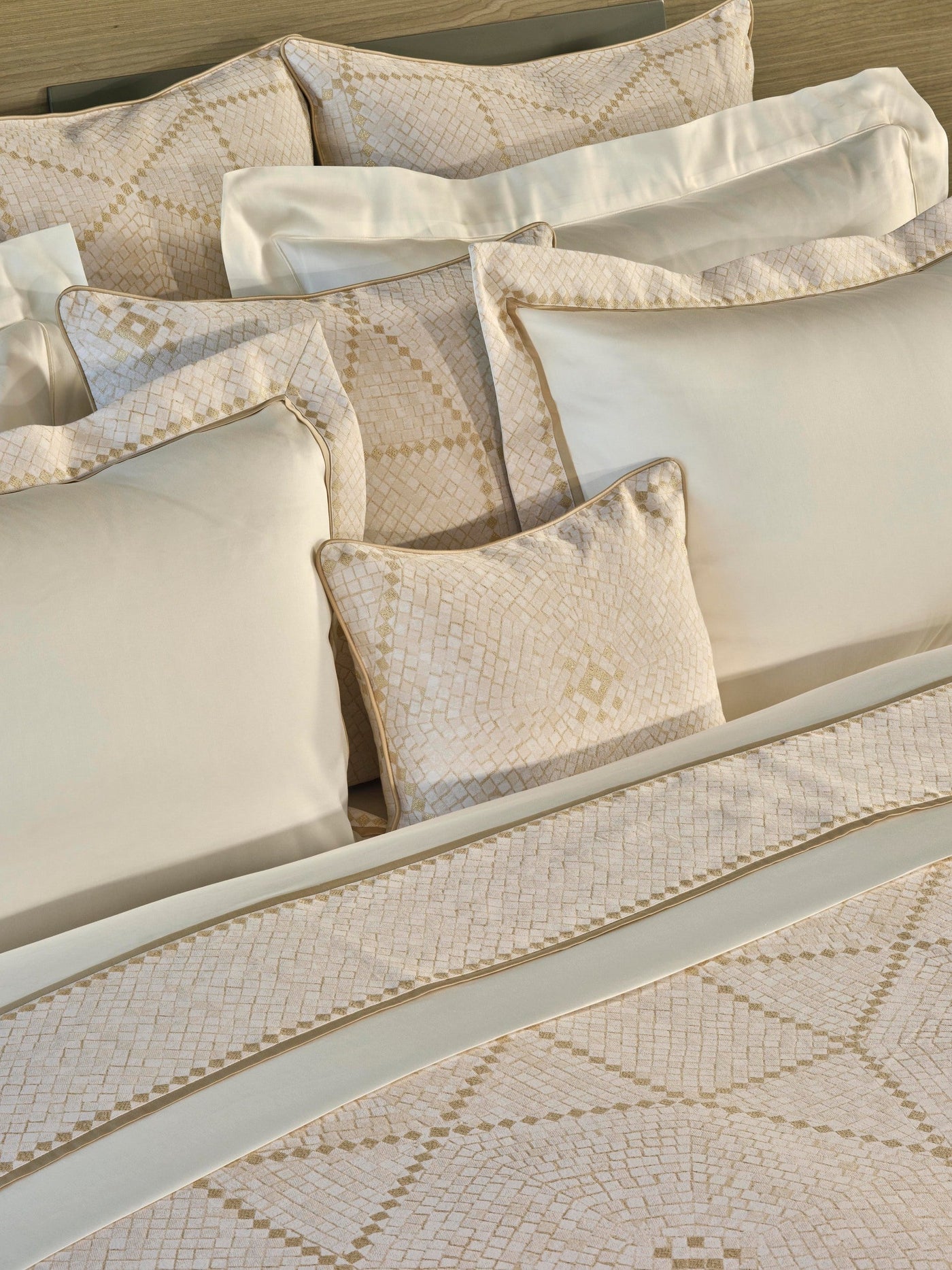 Stacked neutral-toned pillows and bedding feature a textured geometric pattern with subtle gold accents and piped edging, featuring Kerylos Pillowcase by Celso de Lemos available at Linen Society.