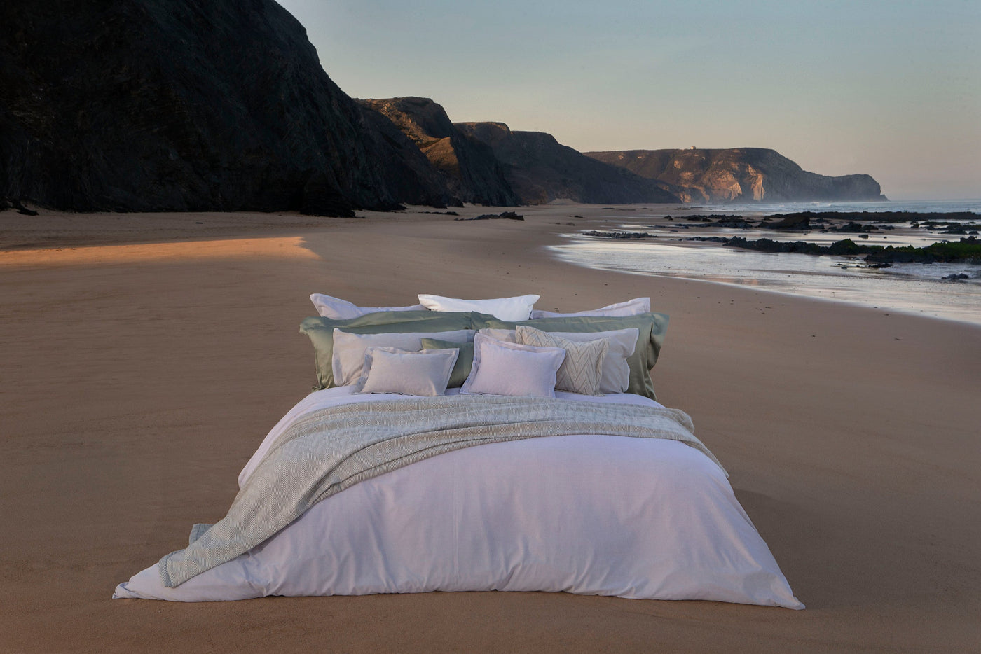 A bed with white and pale green linens is set up on a beach with dramatic cliffs in the background, featuring Kroco Pillowcases by Celso de Lemos available at Linen Society.