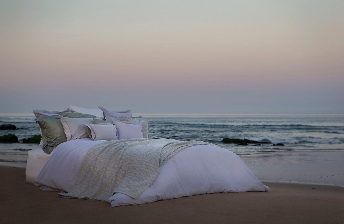 A bed with white and light green bedding is set up on a beach facing the ocean at dusk. featuring Kroco Pillowcase by Celso de Lemos available at Linen Society.