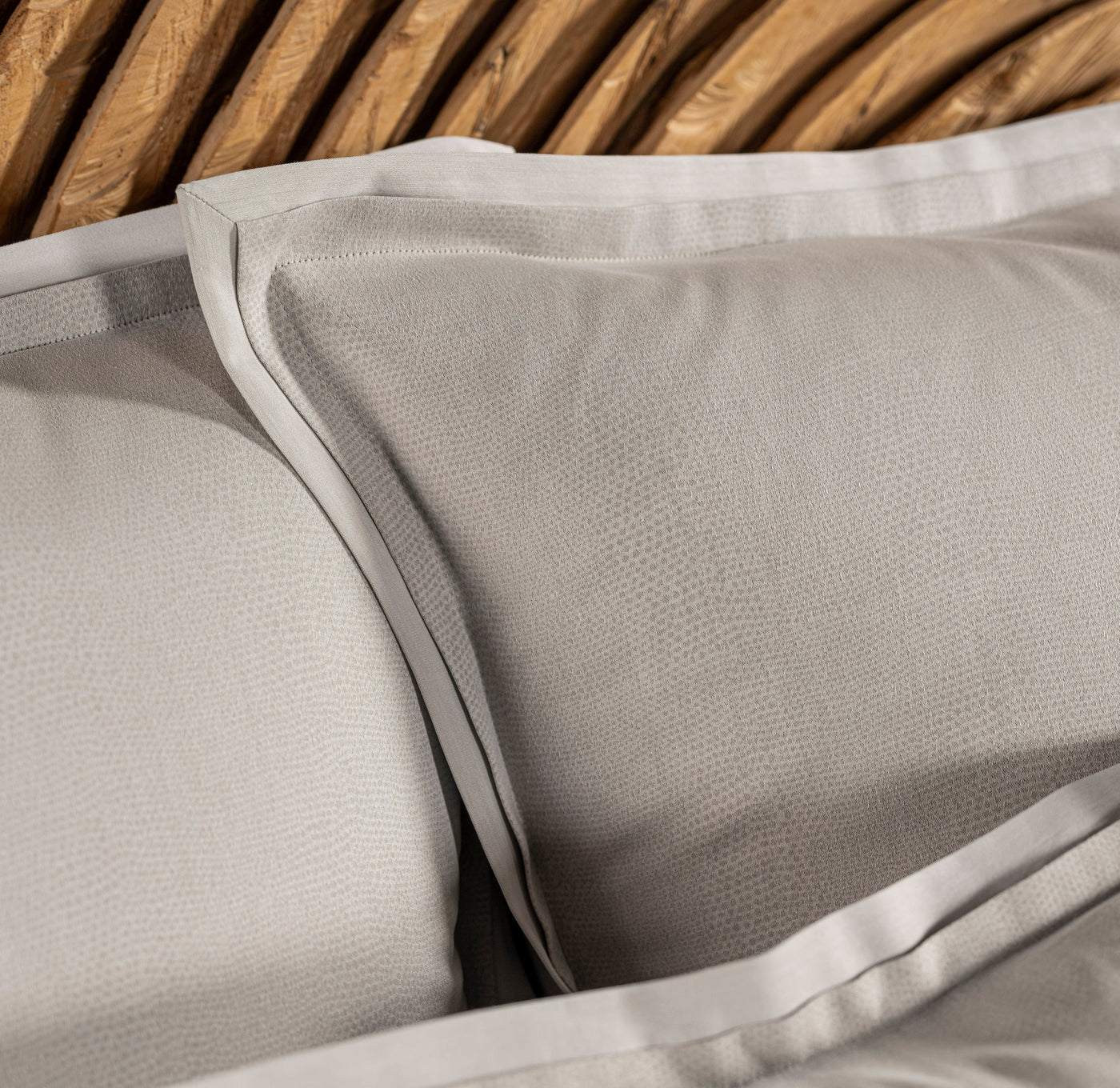 Two light grey pillowcases with a subtle textured pattern are layered against a rustic wooden headboard. Featuring Kroco Pillowcase by Celso de Lemos available at Linen Society.
