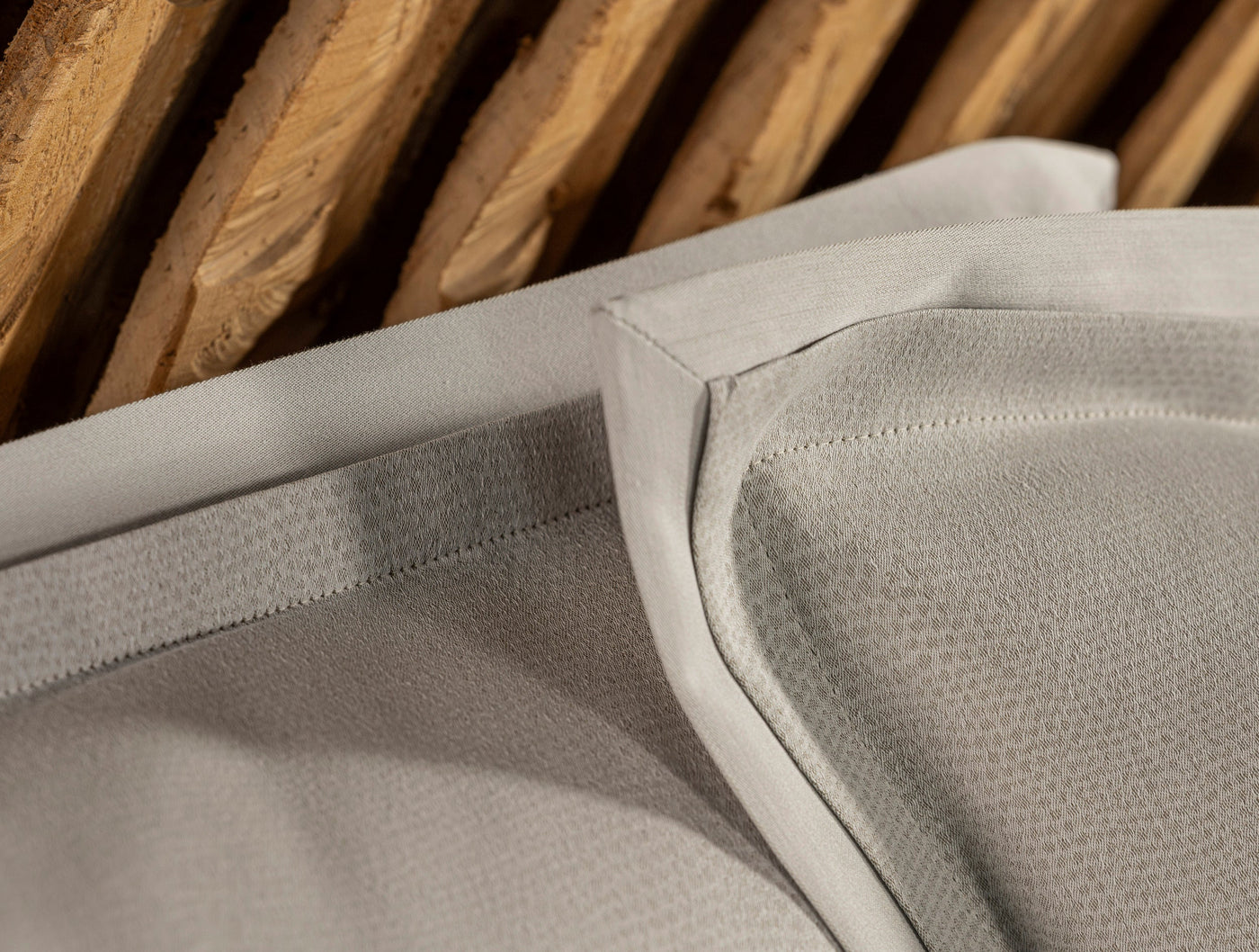 A close-up of textured, light grey pillowcases folded with wooden slats in the background, featuring Kroco Pillowcase by Celso de Lemos available at Linen Society.