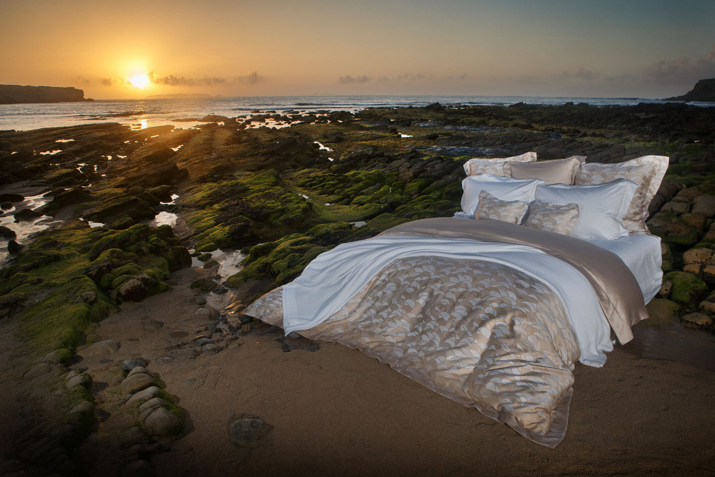 A bed is set up on a rocky, moss-covered shore as the sun sets over the ocean, featuring the Loulou Pillowcase by Celso de Lemos available at Linen Society.