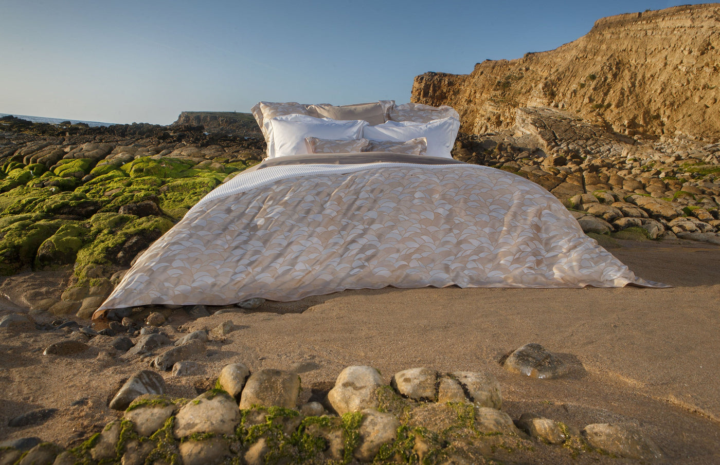 A bed is set up on a sandy beach with moss-covered rocks in the foreground and a cliff in the background. Featuring Loulou Pillowcase by Celso de Lemos available at Linen Society.