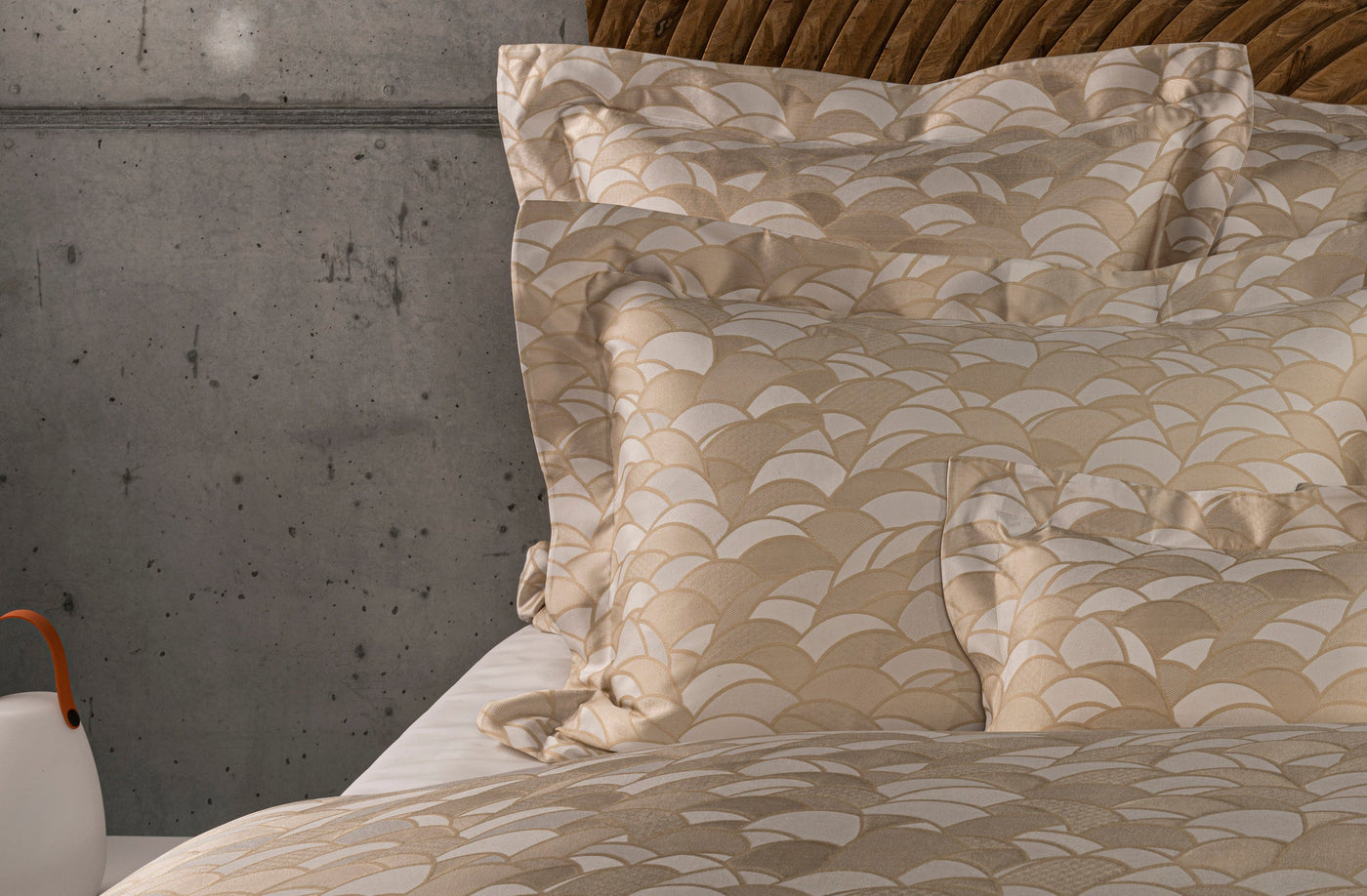 Close-up of a bed with beige and cream patterned pillowcases featuring an art deco fan motif, set against a concrete wall and wooden headboard, featuring Loulou Pillowcase by Celso de Lemos available at Linen Society.