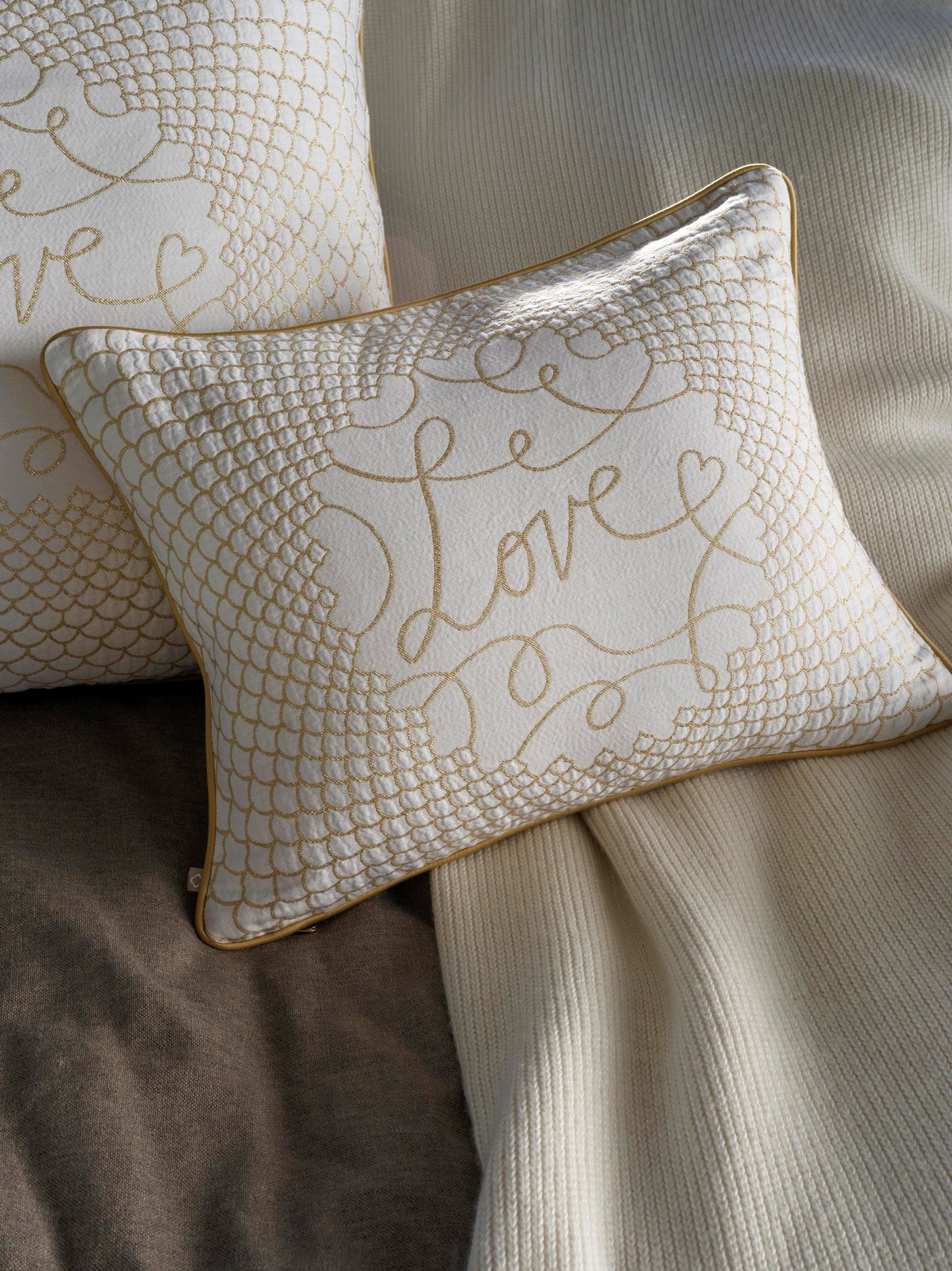 White pillow with gold embroidered "Love" script and intricate scale pattern, featuring Love Pillow Cover by Celso de Lemos available at Linen Society.