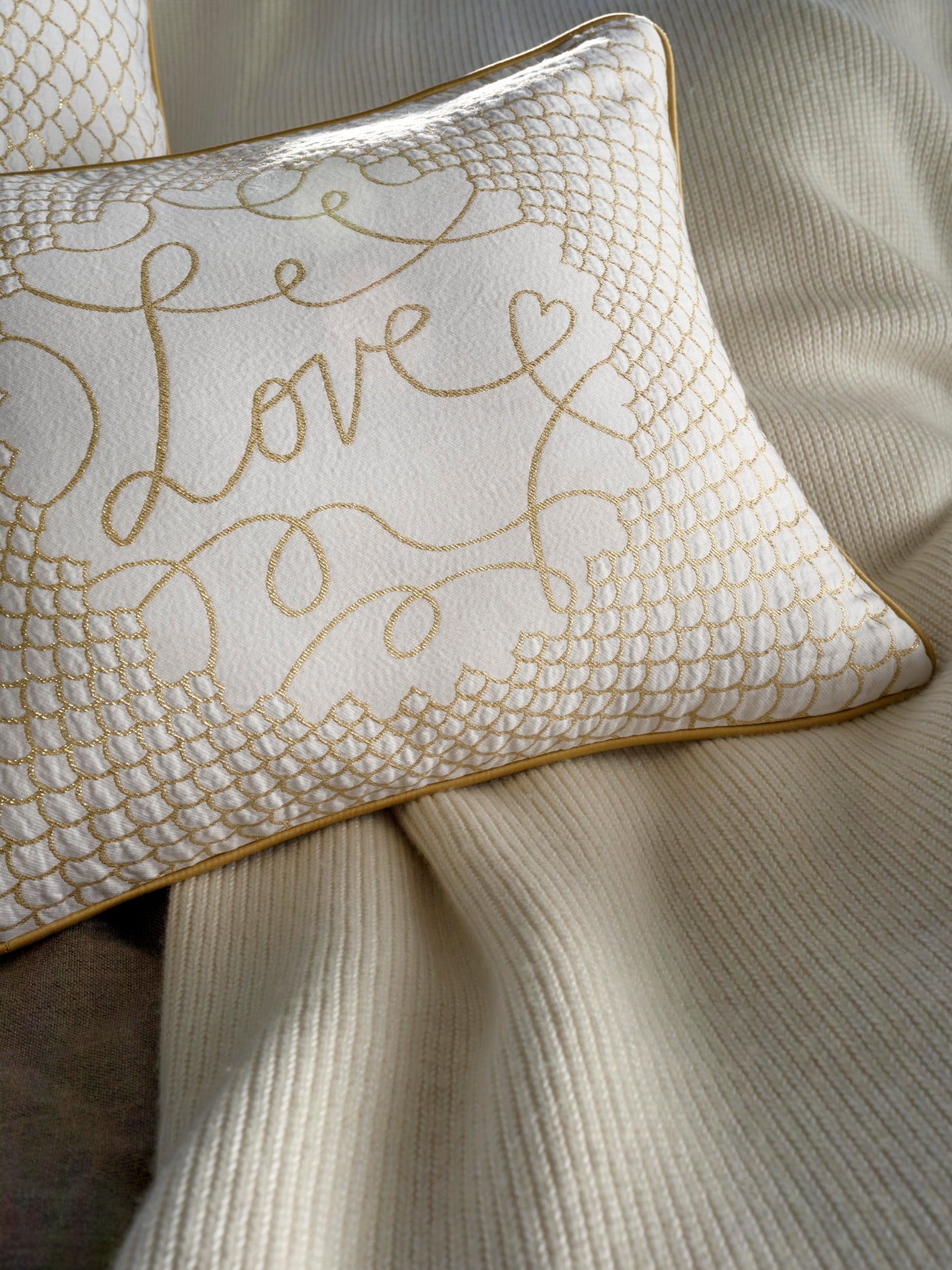 A white pillow with gold embroidery of the word "Love" and a delicate scalloped border lies on a cream-colored textured throw, featuring Love Pillow Cover by Celso de Lemos available at Linen Society.