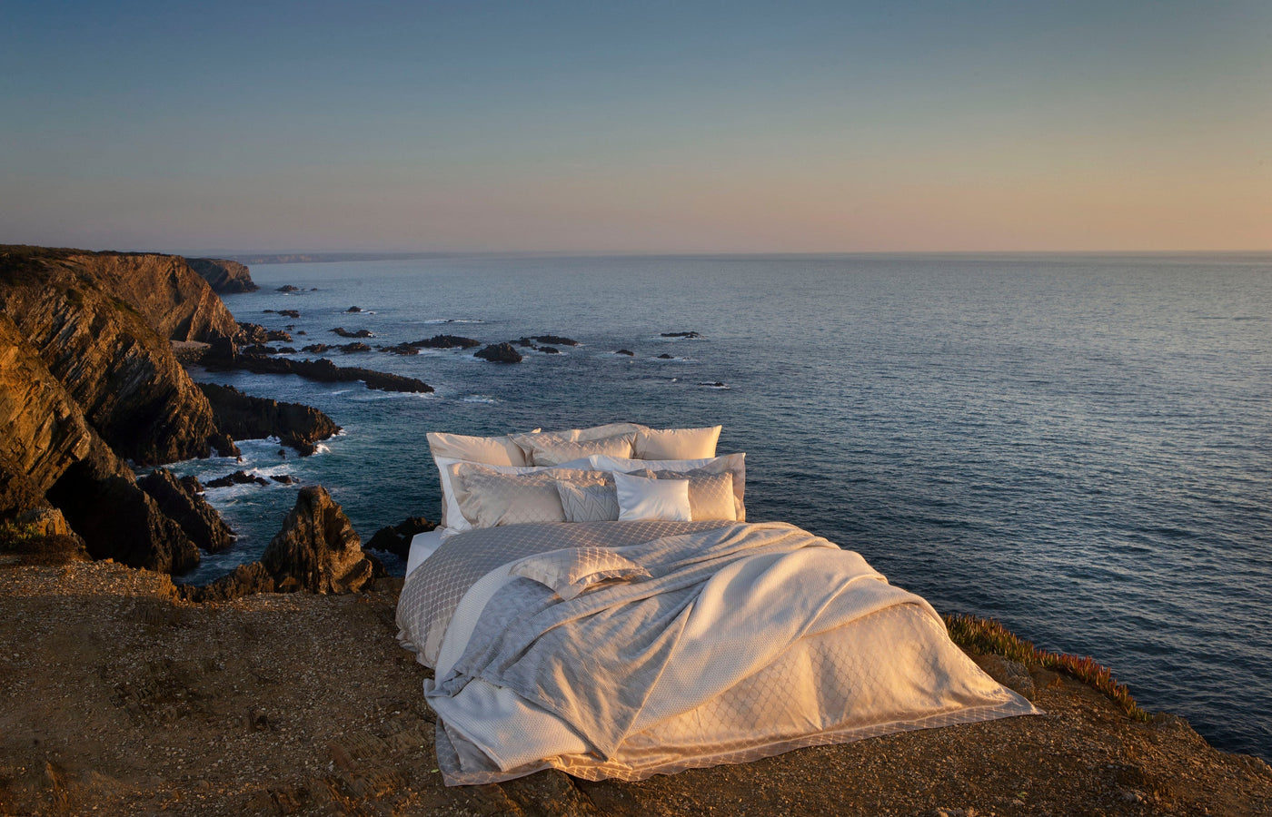 A bed is styled on a cliff overlooking the ocean at sunset, featuring the Lua Pillowcase by Celso de Lemos available at Linen Society.