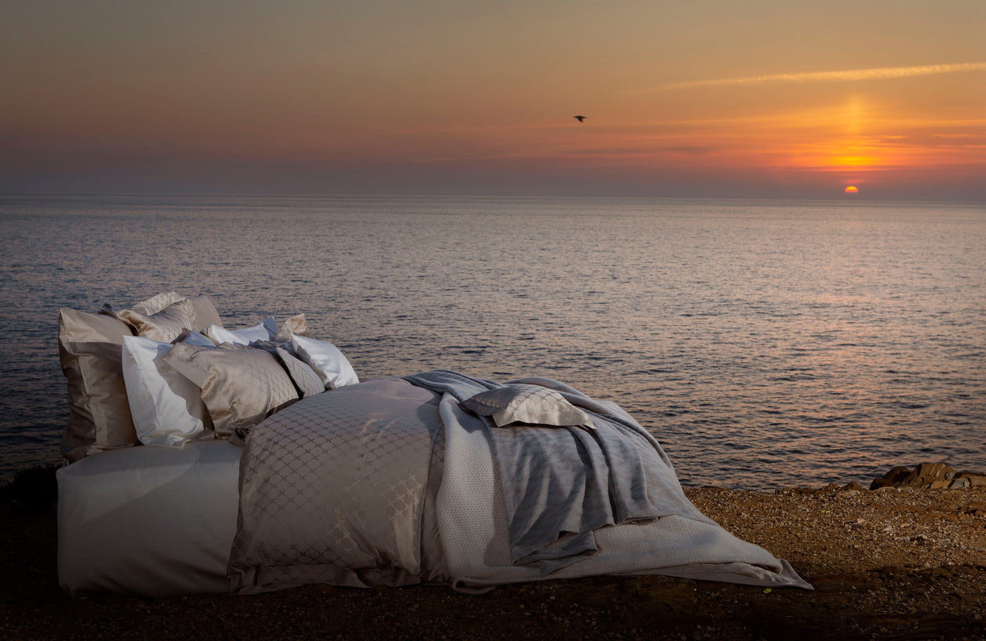 A bed with a white and tan patterned duvet and pillows is set up on a rocky shore facing the ocean at sunset, featuring Lua Pillowcase by Celso de Lemos available at Linen Society.