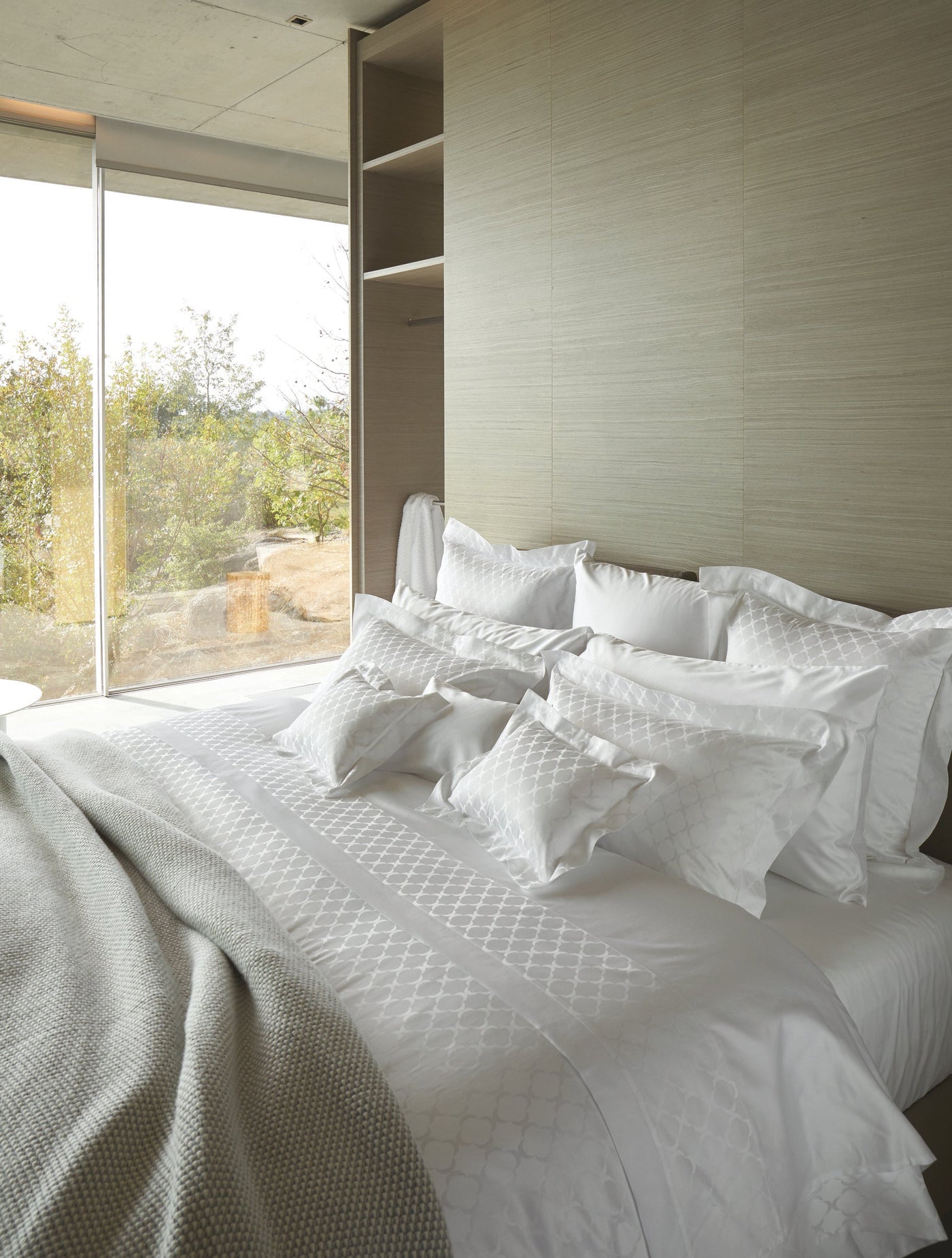 A neatly made bed with white bedding featuring an embossed geometric pattern, a light grey throw, and large windows overlooking greenery, featuring Lua Pillowcase by Celso de Lemos available at Linen Society.