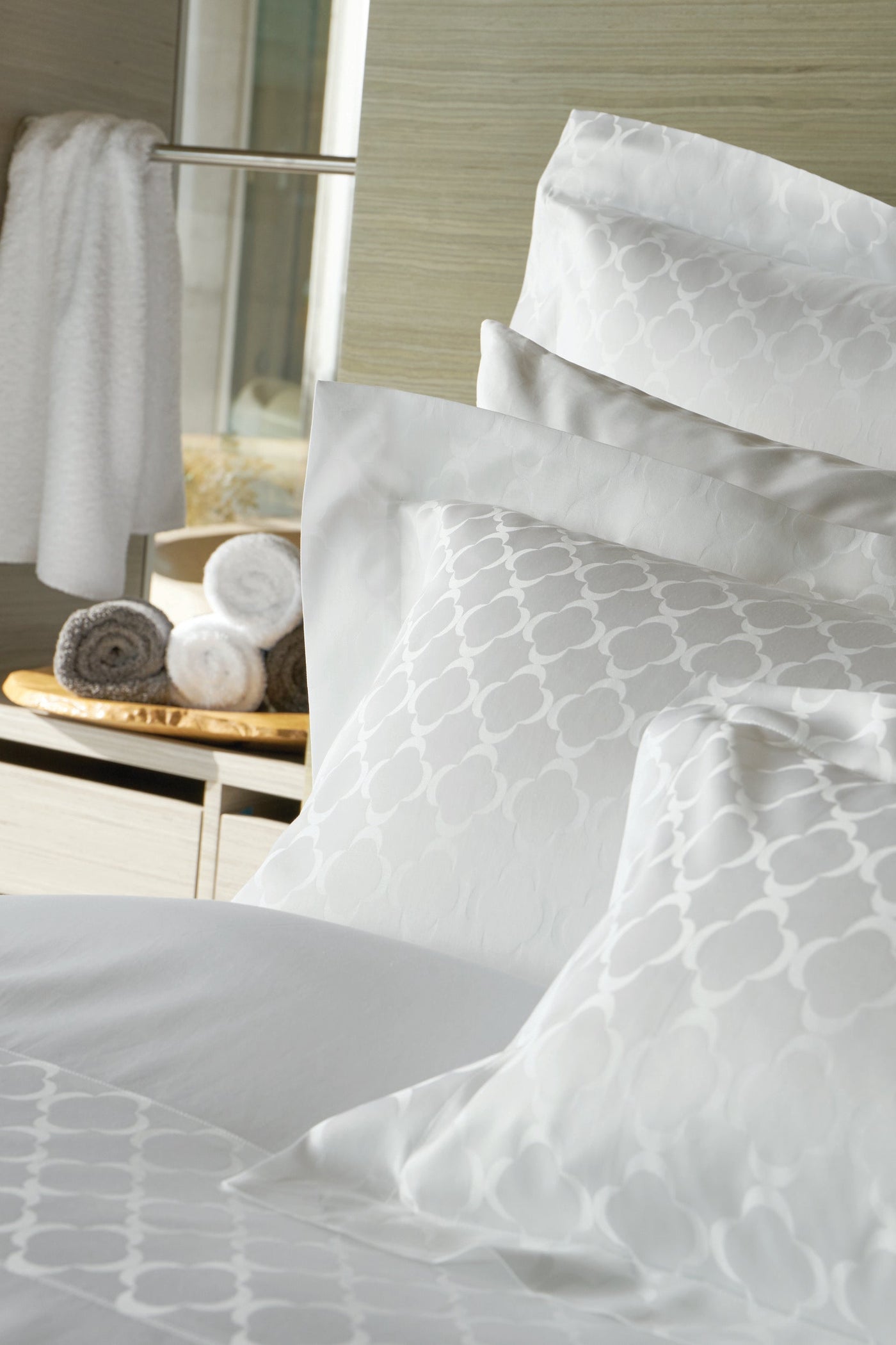 Stacked white pillows with a subtle quatrefoil pattern and a white towel hanging in the background, featuring Lua Pillowcase by Celso de Lemos available at Linen Society.
