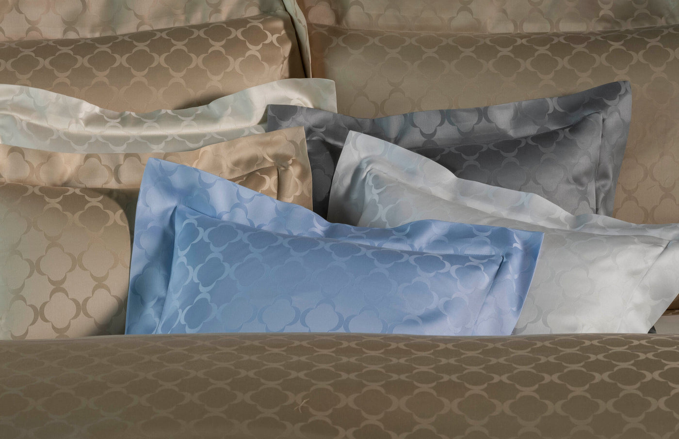 A stack of folded pillowcases in various neutral and pastel colors, featuring a subtle quatrefoil pattern. featuring Lua Pillowcase by Celso de Lemos available at Linen Society.