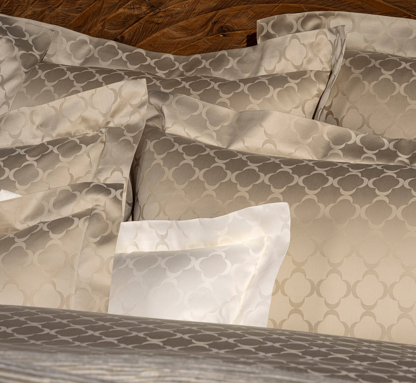 A close-up view of stacked linen pillows and bedding featuring a subtle quatrefoil pattern in neutral tones, including cream and taupe, on a wooden headboard. featuring Lua Pillowcase by Celso de Lemos available at Linen Society.