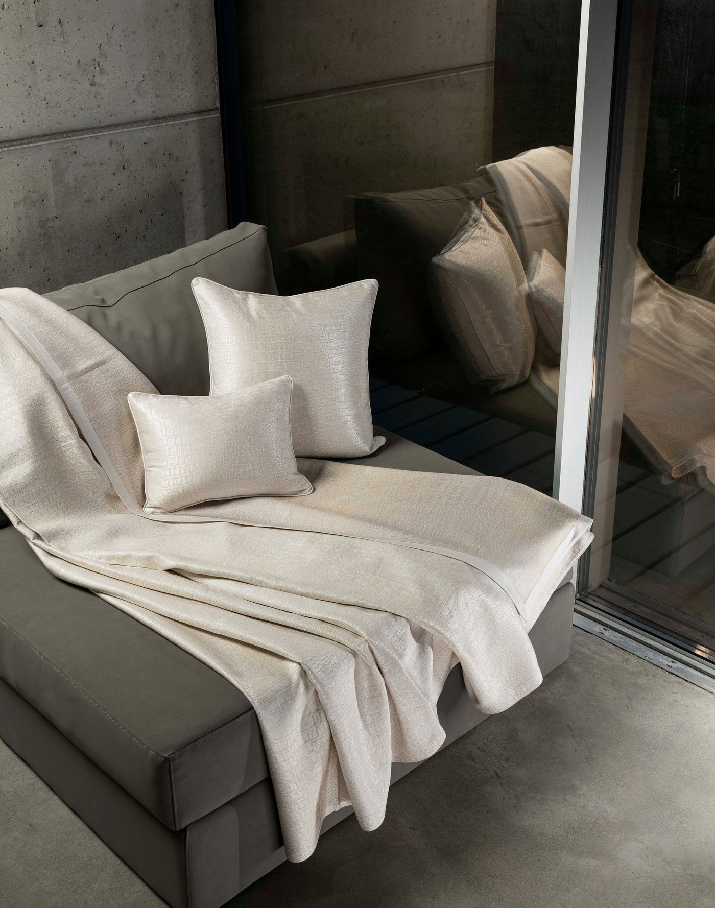 Two light-colored throw pillows with a textured, embossed pattern are placed on a sofa, draped with a matching throw, featuring Luxe Pillow Cover by Celso de Lemos available at Linen Society.