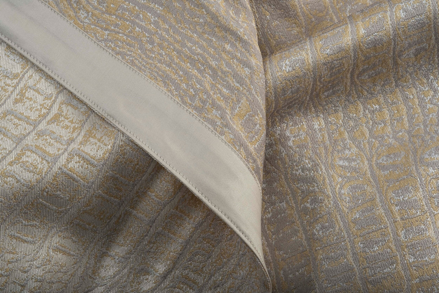 A textured fabric with a geometric pattern in gold and taupe, folded to reveal a plain taupe band, featuring Luxe Pillow Cover by Celso de Lemos available at Linen Society.