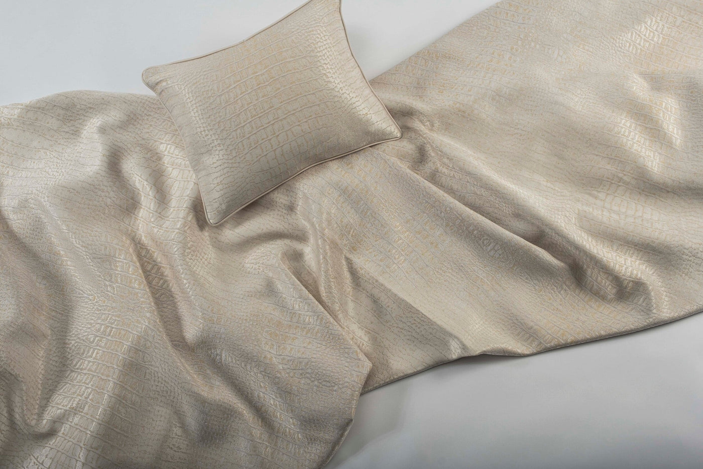 A beige pillow rests on a textured, shimmering fabric with a crocodile-like pattern, featuring Luxe Pillow Cover by Celso de Lemos available at Linen Society.