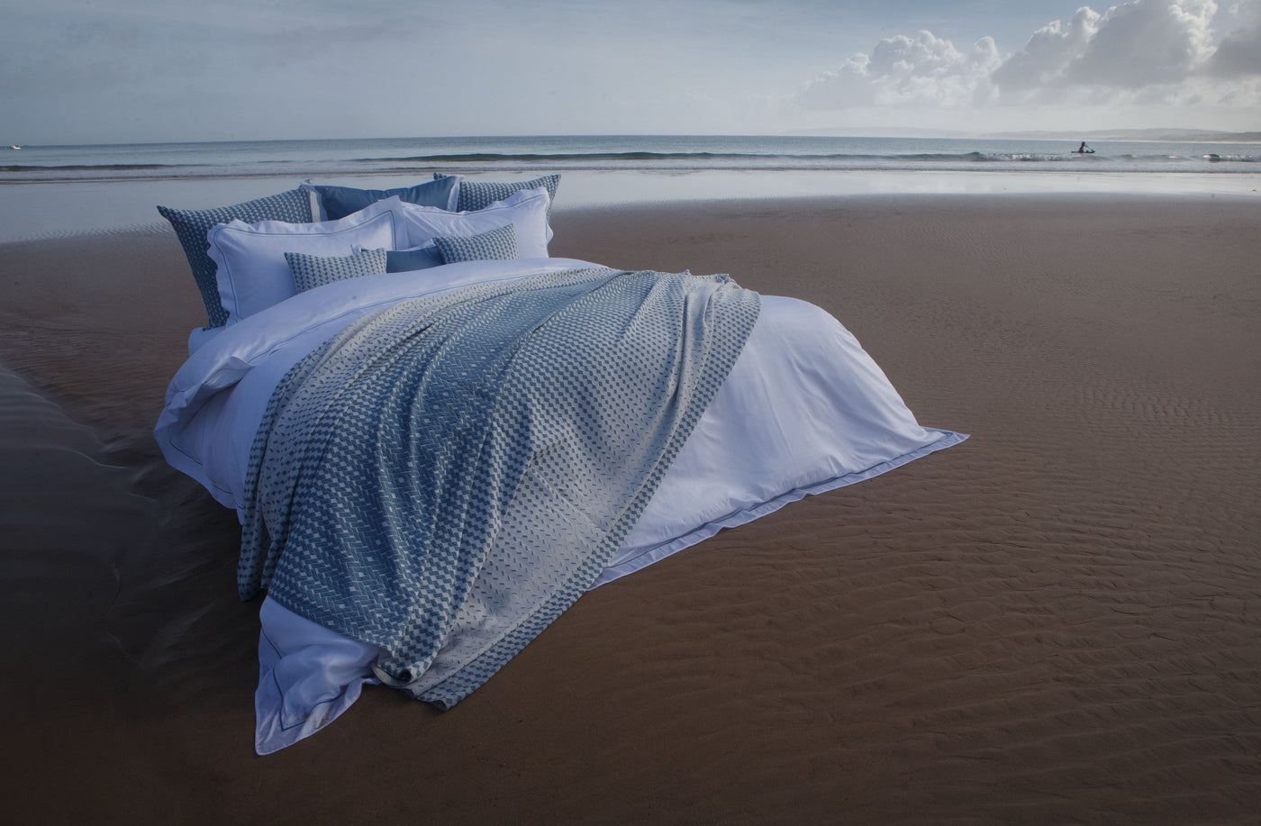 A bed with white bedding and a blue textured throw is set on a sandy beach overlooking the ocean, featuring Marbella Pillow Cover by Celso de Lemos available at Linen Society.