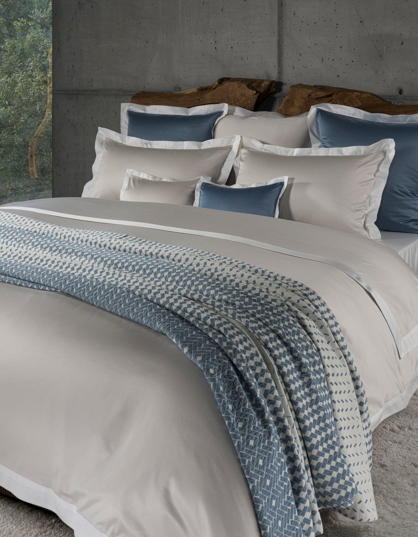 A neatly made bed with a grey duvet and throw, featuring a patterned blanket draped over it, is displayed against a textured concrete wall, featuring Marbella Pillow Cover by Celso de Lemos available at Linen Society.