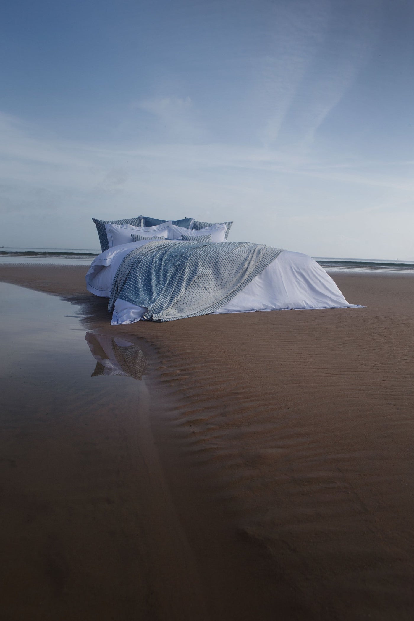 A bed with white linens and a blue patterned throw is set up on a wet beach near the ocean. featuring Marbella Pillow Cover by Celso de Lemos available at Linen Society.