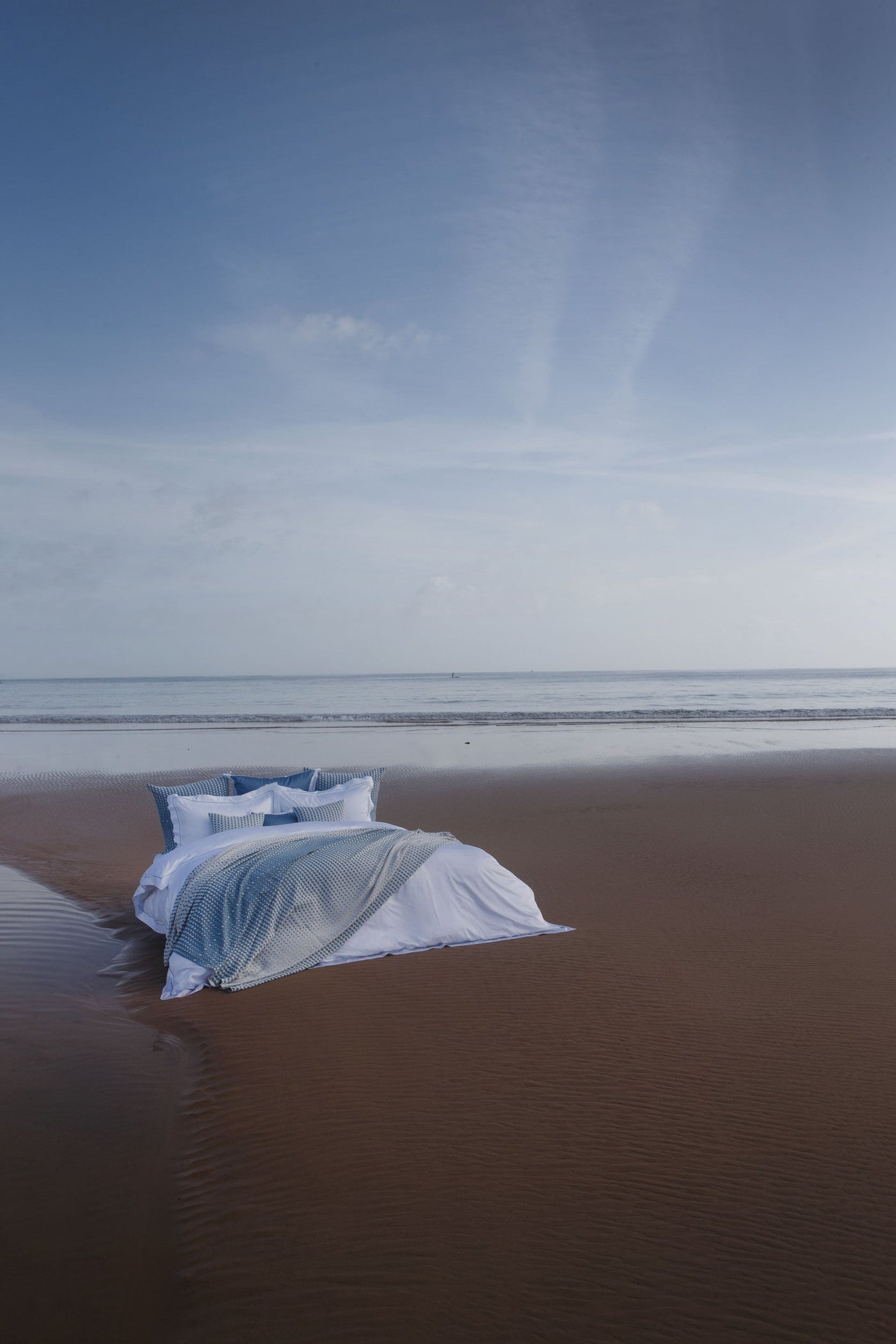 A bed is staged on a sandy beach with the ocean in the background, featuring the Marbella Pillow Cover by Celso de Lemos available at Linen Society.