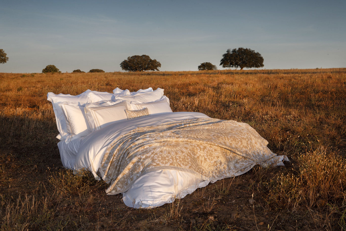 A bed dressed in white linens and a golden-patterned throw sits in a dry, grassy field with trees in the background, featuring Milena Pillow Cover by Celso de Lemos available at Linen Society.