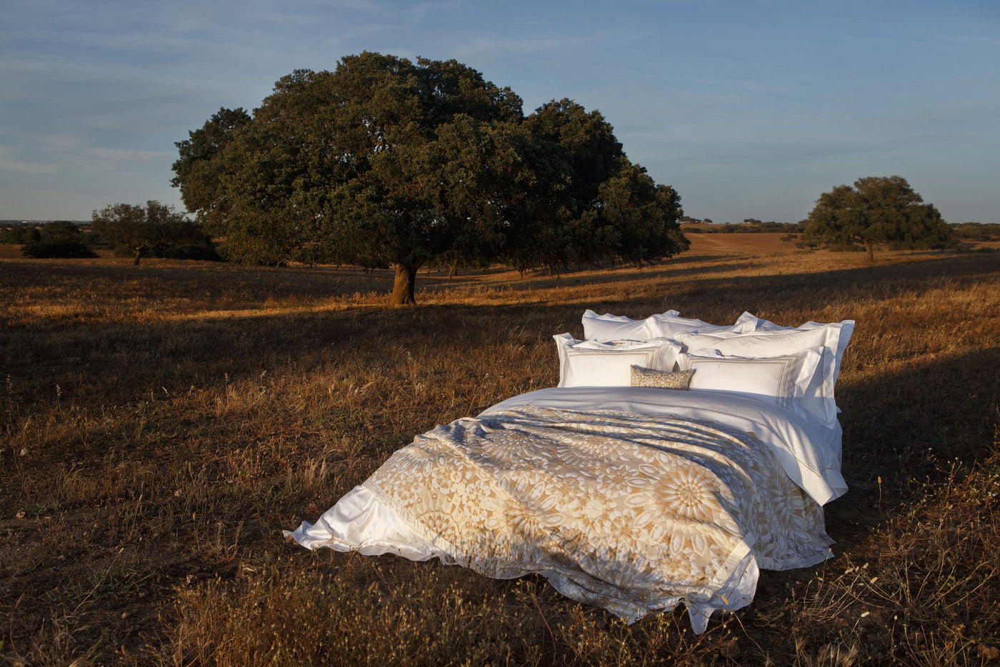 A bed with white linens and a beige and white patterned throw is set in a dry, grassy field under a large oak tree, featuring Milena Pillow Cover by Celso de Lemos available at Linen Society.