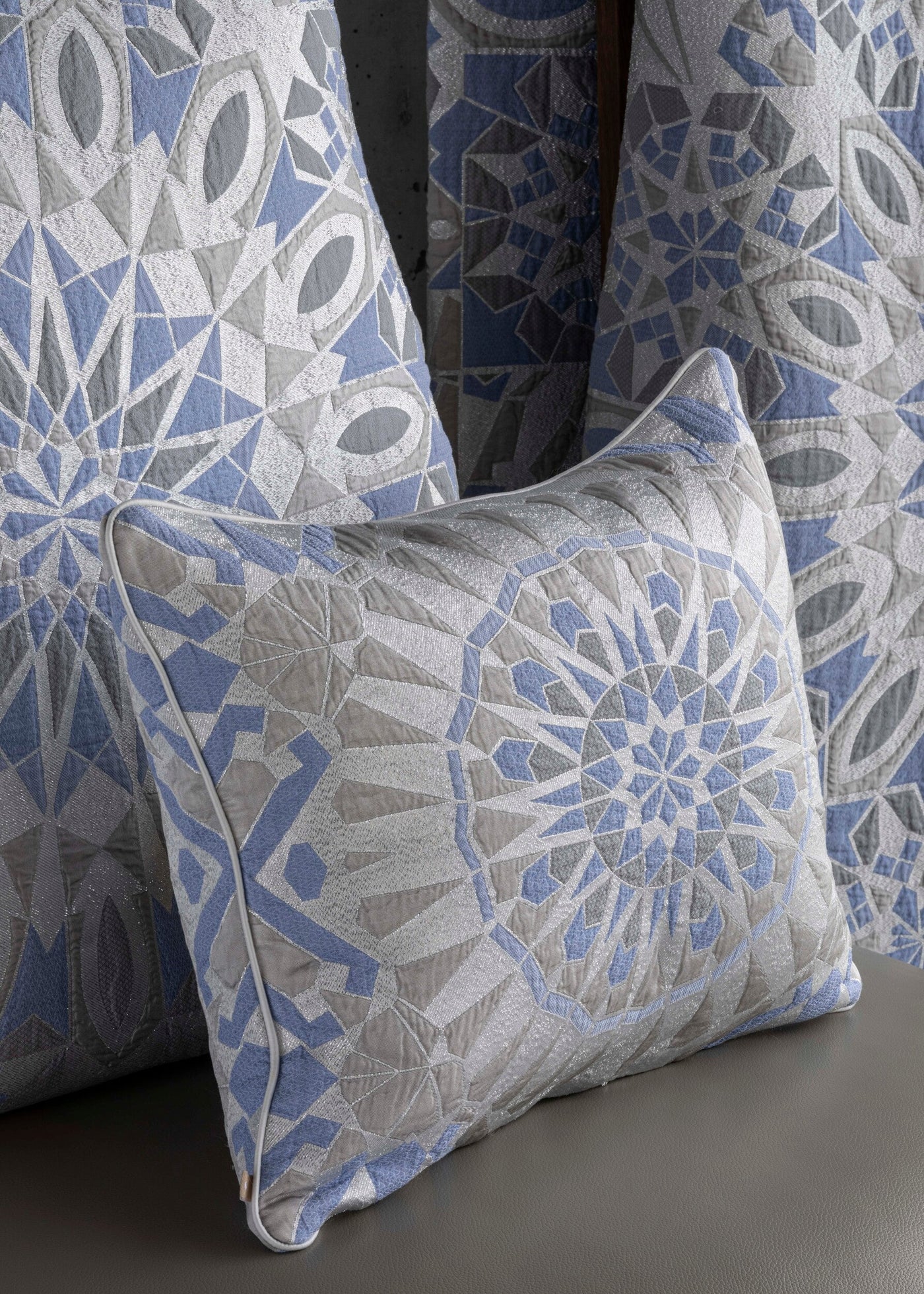 A quilted pillow with a geometric pattern in blue, grey, and silver, featuring the Milena Pillow Cover by Celso de Lemos available at Linen Society.
