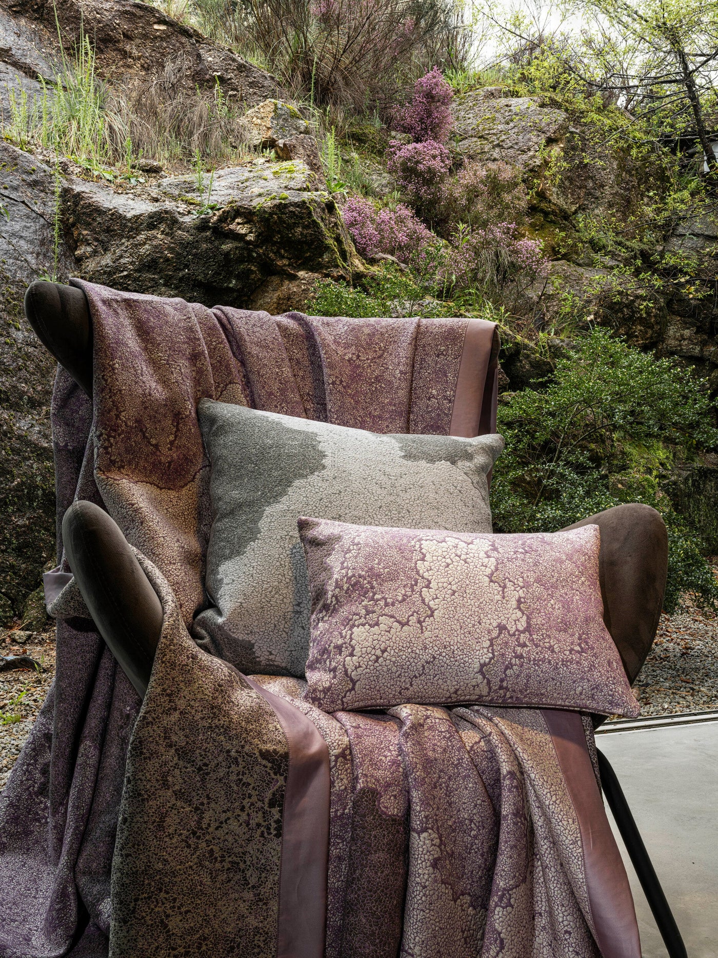 Two textured, patterned pillow covers in grey and dusty rose are placed on an armchair draped with a matching throw blanket, featuring the Mundo Pillow Cover by Celso de Lemos available at Linen Society.