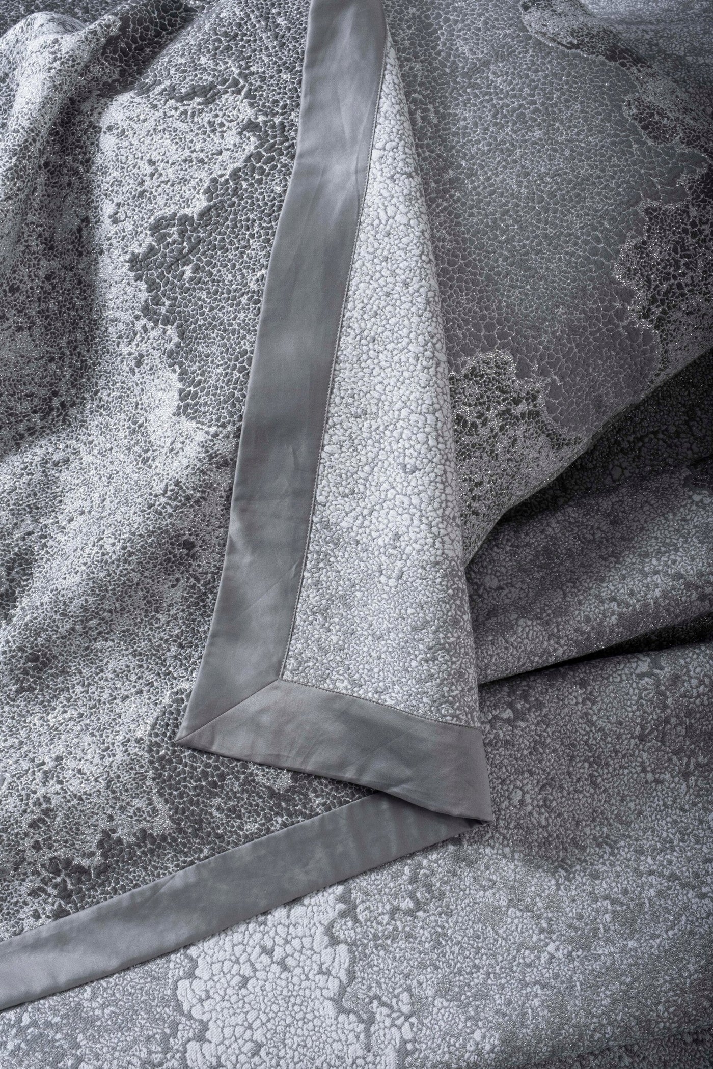 Close-up of a grey pillow cover with a textured, crackled pattern and a smooth grey satin trim, featuring Mundo Pillow Cover by Celso de Lemos available at Linen Society.