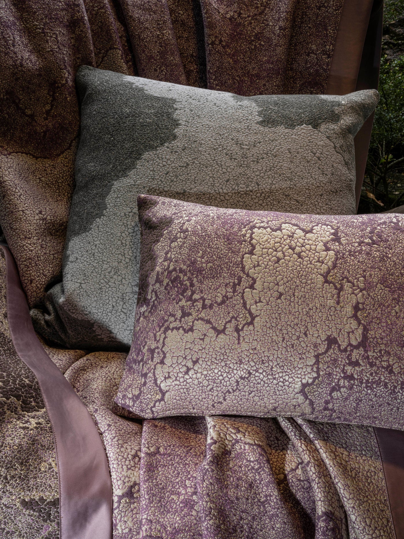 Two textured pillow covers in purple and grey, draped with fabric featuring an abstract pattern and gold threads, featuring Mundo Pillow Cover by Celso de Lemos available at Linen Society.