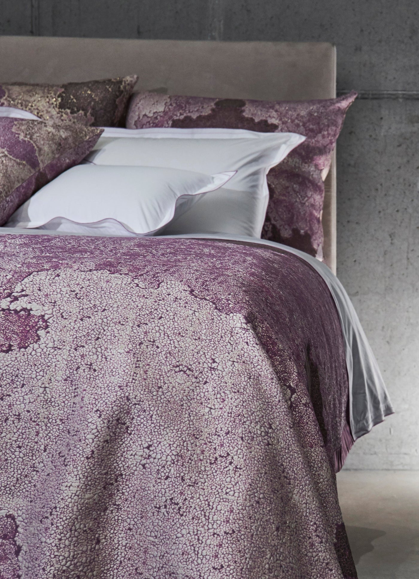 A bed with purple and white patterned pillows and a duvet cover against a concrete wall, featuring Mundo Pillow Cover by Celso de Lemos available at Linen Society.