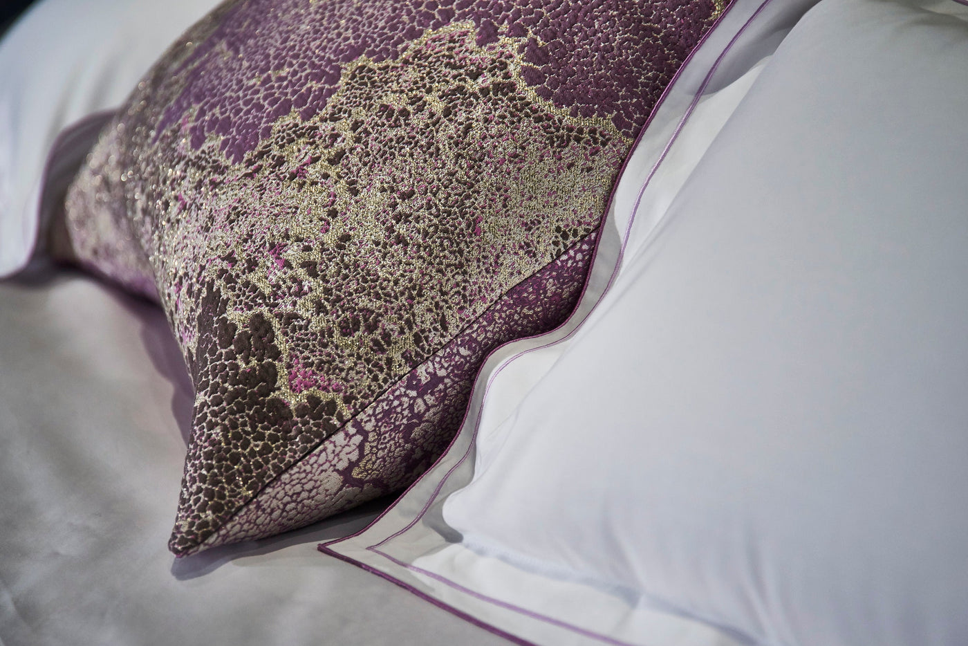 A textured purple pillow with gold accents rests on crisp white bedding featuring a purple trim, offering a luxurious feel. featuring Mundo Pillow Cover by Celso de Lemos available at Linen Society.