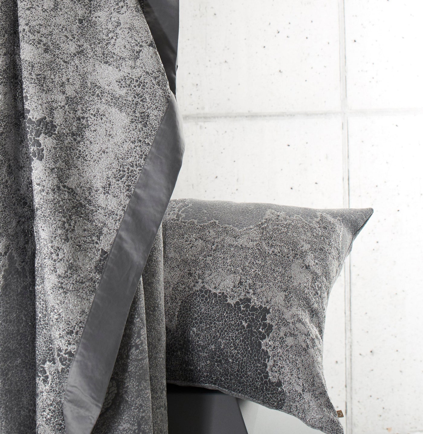 A textured gray throw pillow rests against a draped fabric with a similar textured pattern and a satin trim. featuring Mundo Pillow Cover by Celso de Lemos available at Linen Society.