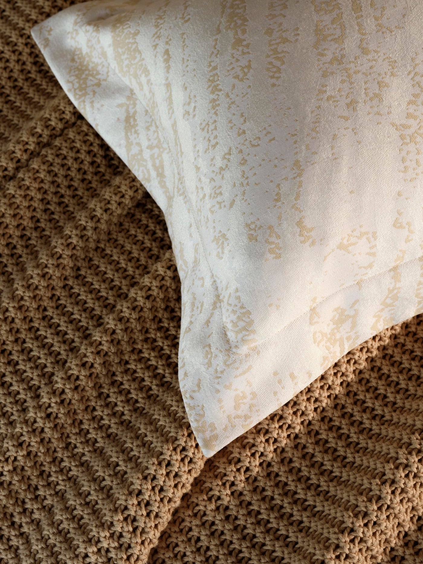 Close-up of Neve pillowcase corner showing ivory cotton with champagne speckled print against a caramel knit blanket.