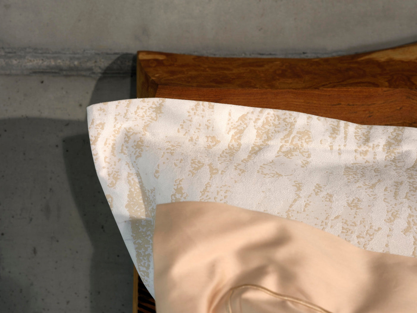 Close-up of ivory and gold jacquard fabric draped over a wooden headboard with a champagne pillowcase below.