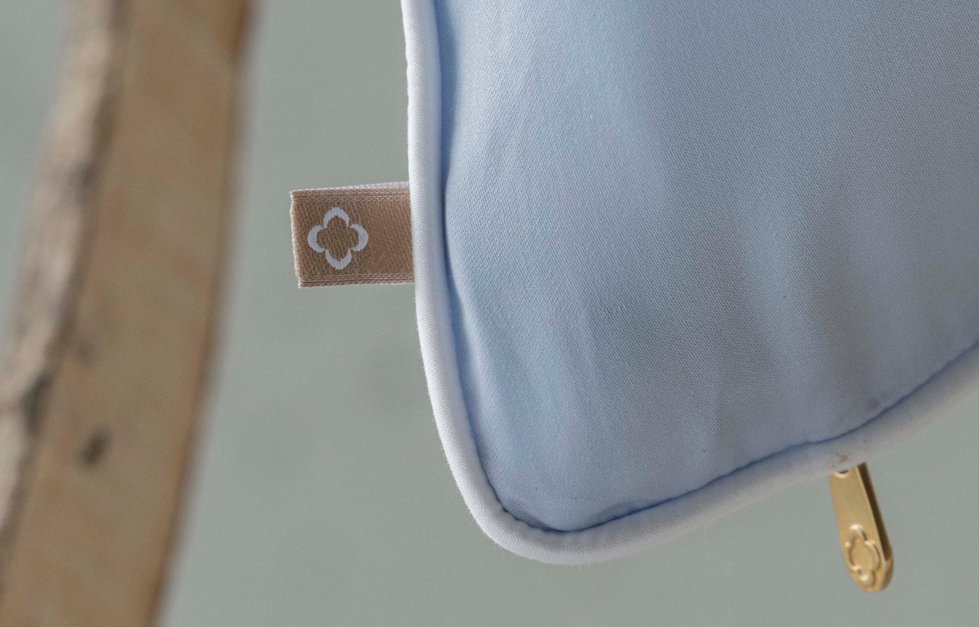 Close-up of a light blue pillow with a white trim and a gold zipper pull featuring the Noa Pillow Cover by Celso de Lemos available at Linen Society.
