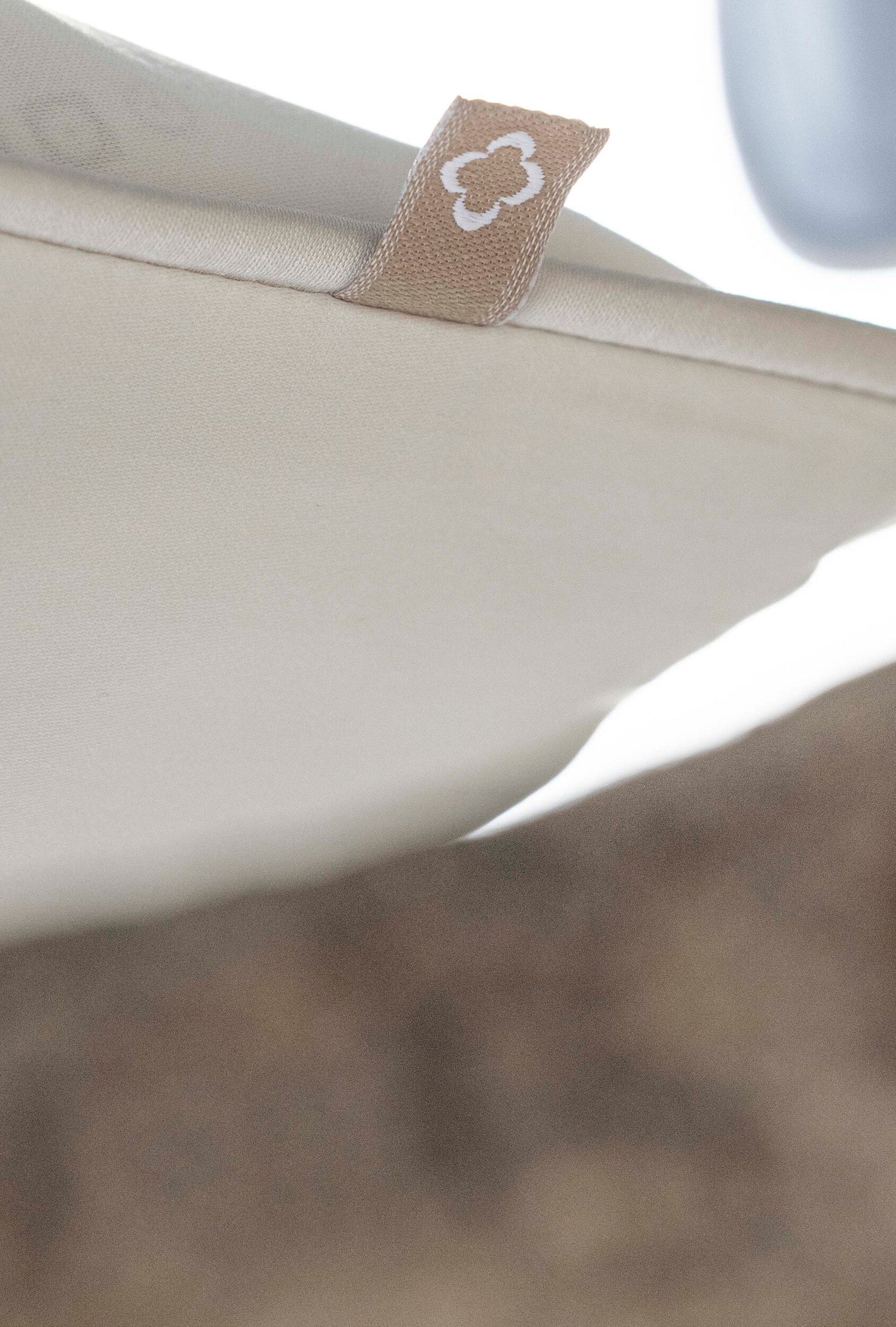 Close-up on a light beige textured fabric with a small tan tag featuring a white quatrefoil pattern, featuring Noa Pillow Cover by Celso de Lemos available at Linen Society.