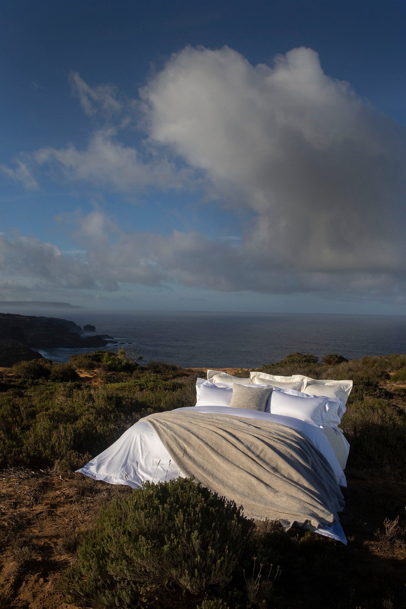 A bed with white linens and a striped textured throw is set up outdoors overlooking the ocean. featuring Panama Pillow Cover by Celso de Lemos available at Linen Society.